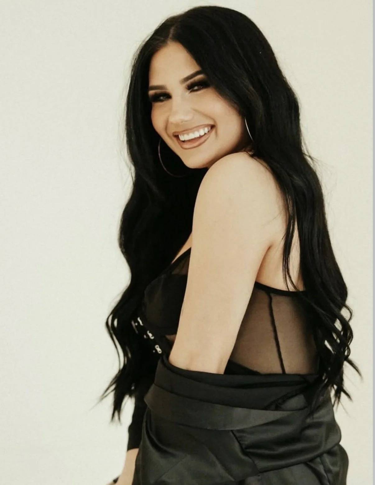 A woman with long black hair, wearing large hoop earrings, smile, black sheer top, and black leather pants, standing against a plain, light-colored wall.