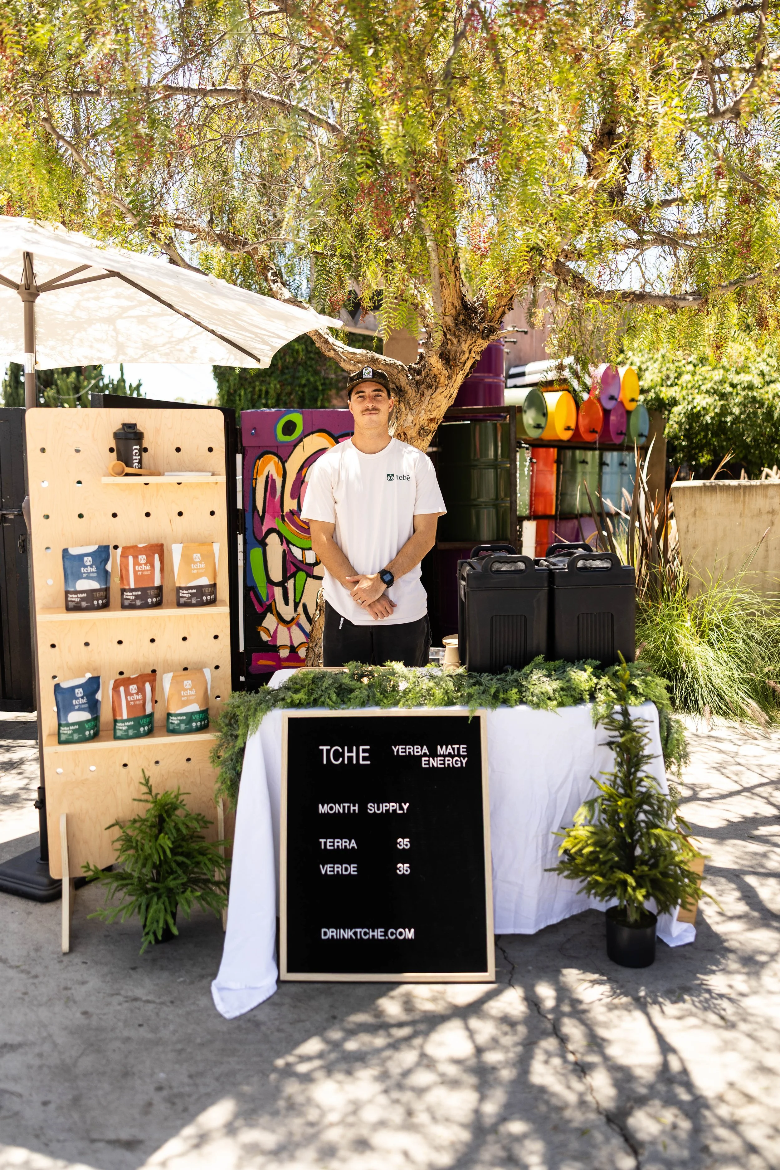 A man in a white t-shirt and cap standing in front of a table with herbal tea products and a blackboard sign at an outdoor market stall, with a large tree and colorful drums in the background.