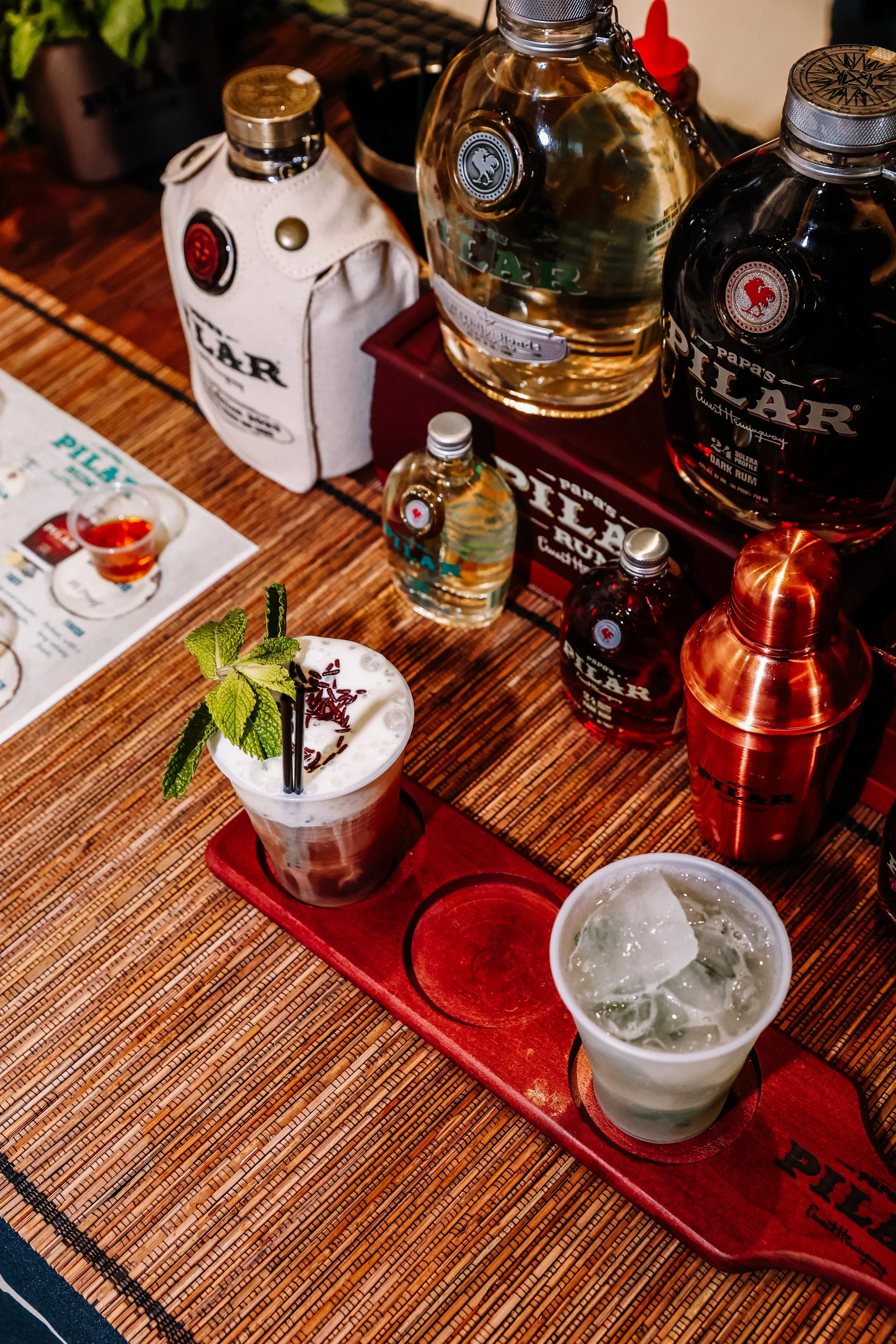 Two cocktails served on a red wooden tray on a woven placemat, with bottles of alcohol in the background.