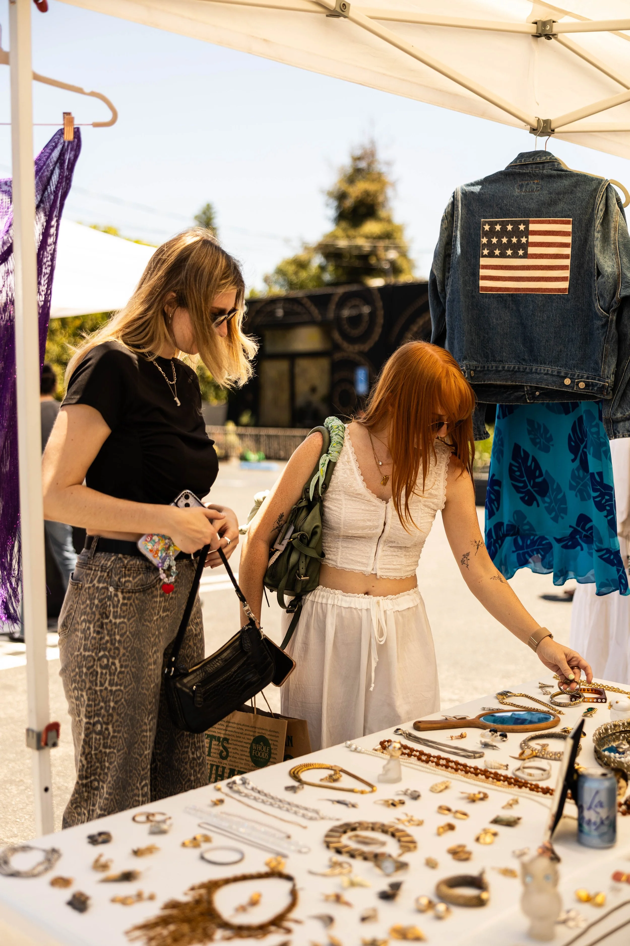People browsing jewelry at an outdoor market stall, with vintage denim jacket featuring an American flag patch on display.