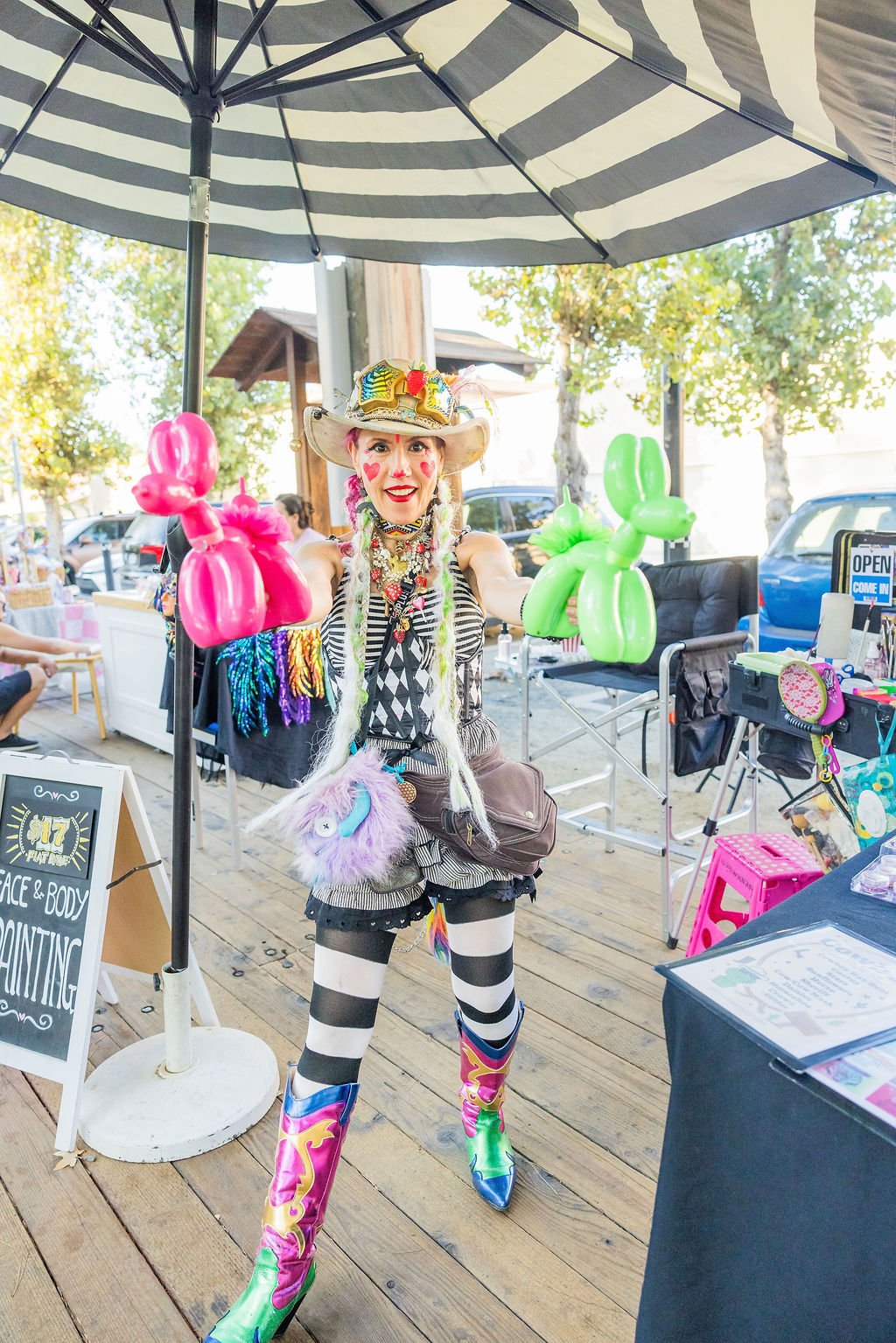 A woman in colorful, whimsical costume holding pink and green balloon animals, standing under a striped parasol at an outdoor event.