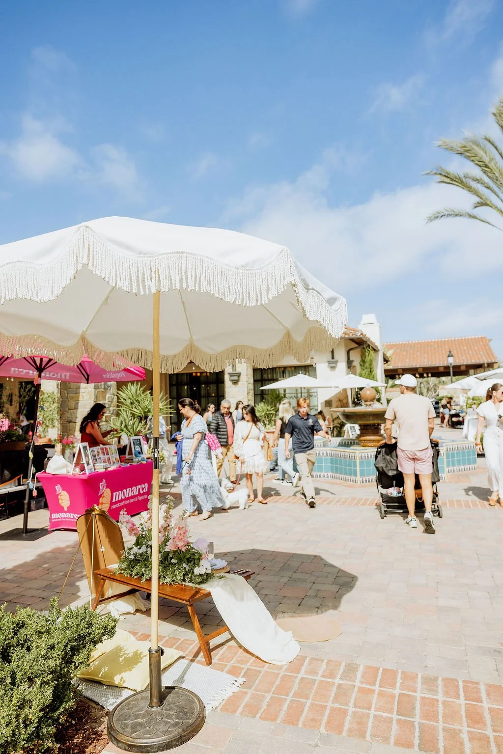Outdoor shopping area with people walking, vendors, and a water fountain, under umbrellas on a sunny day.