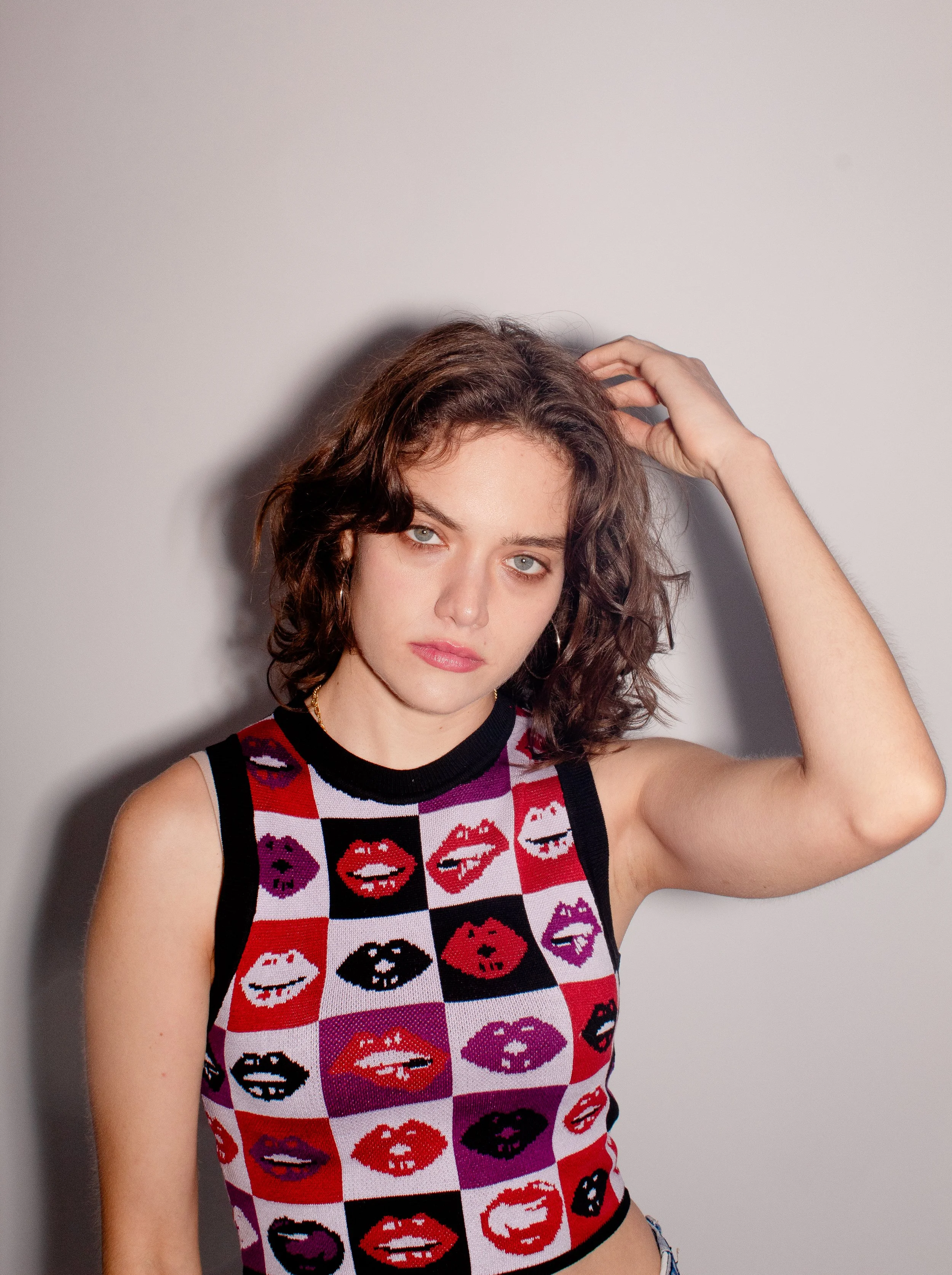 A woman with wavy brown hair and blue eyes wearing a sleeveless top with a colorful lip print pattern, standing against a plain light gray wall.