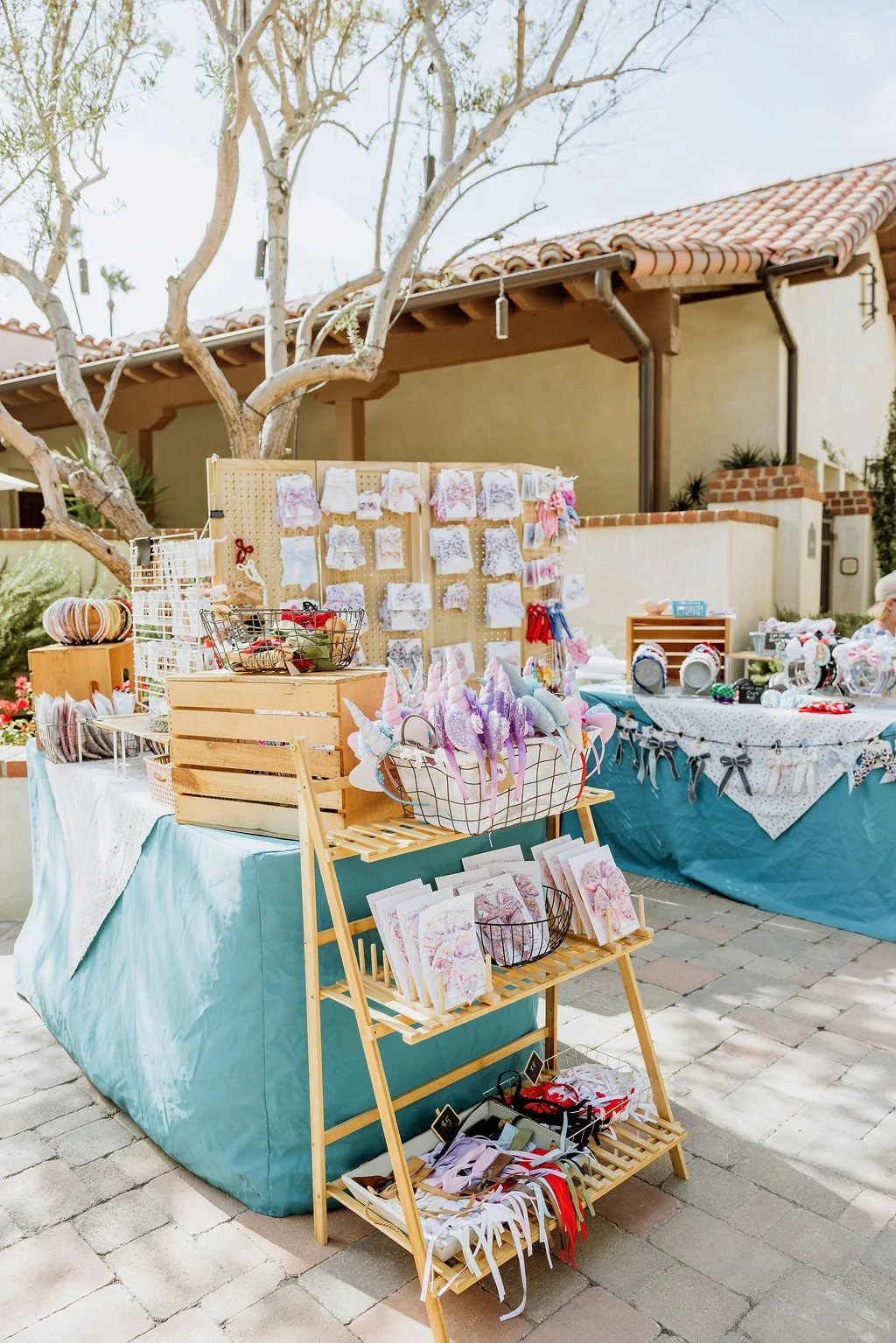 Outdoor craft market stall with pastel-colored unicorn headbands, hair accessories, and decorative items displayed on tables and a wooden rack in a sunny backyard.