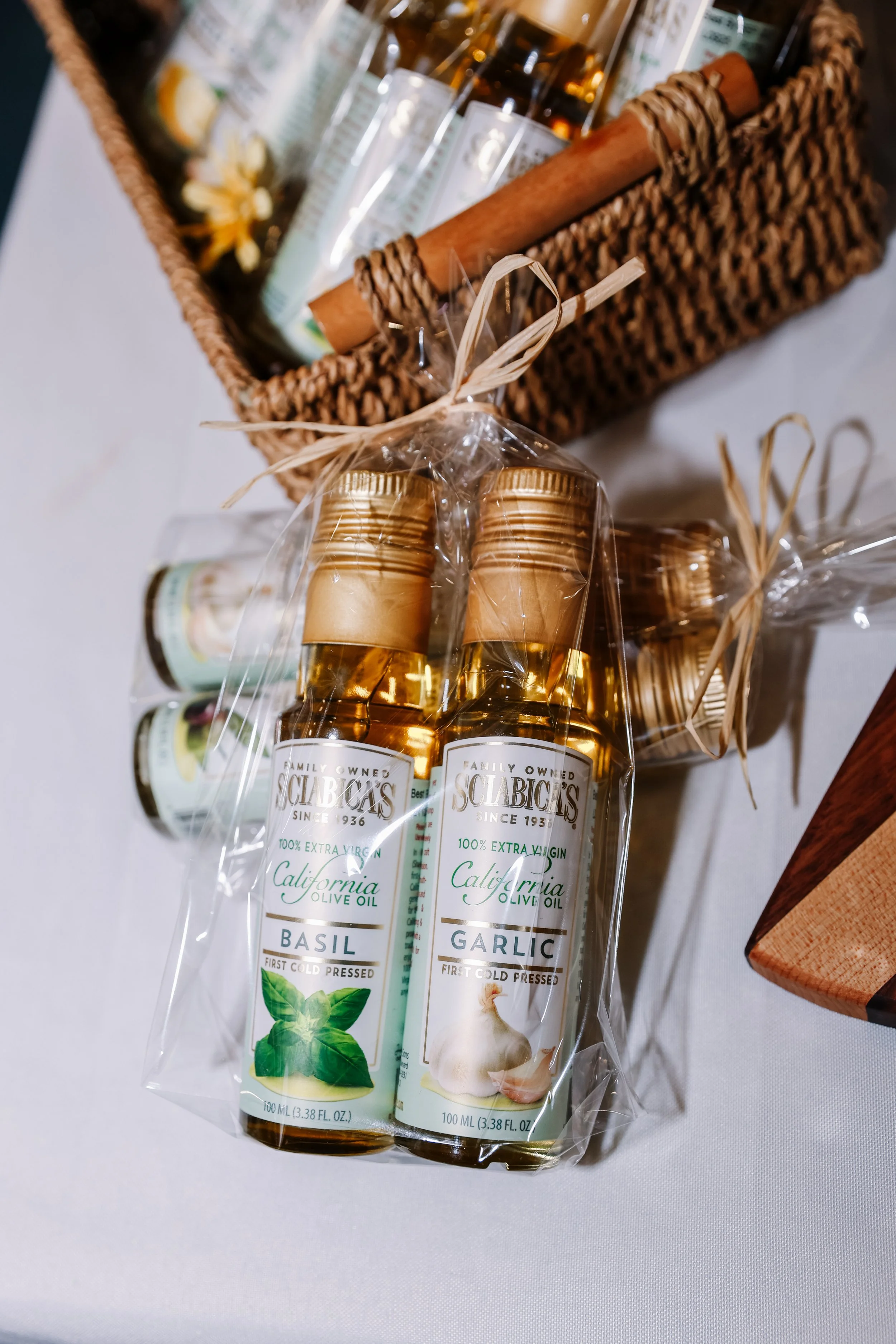 Three bottles of California olive oil, labeled basil, garlic, and one partially visible, packaged in clear plastic with a tied ribbon. A woven basket in the background contains various items, including glass bottles and what appears to be a stick or 