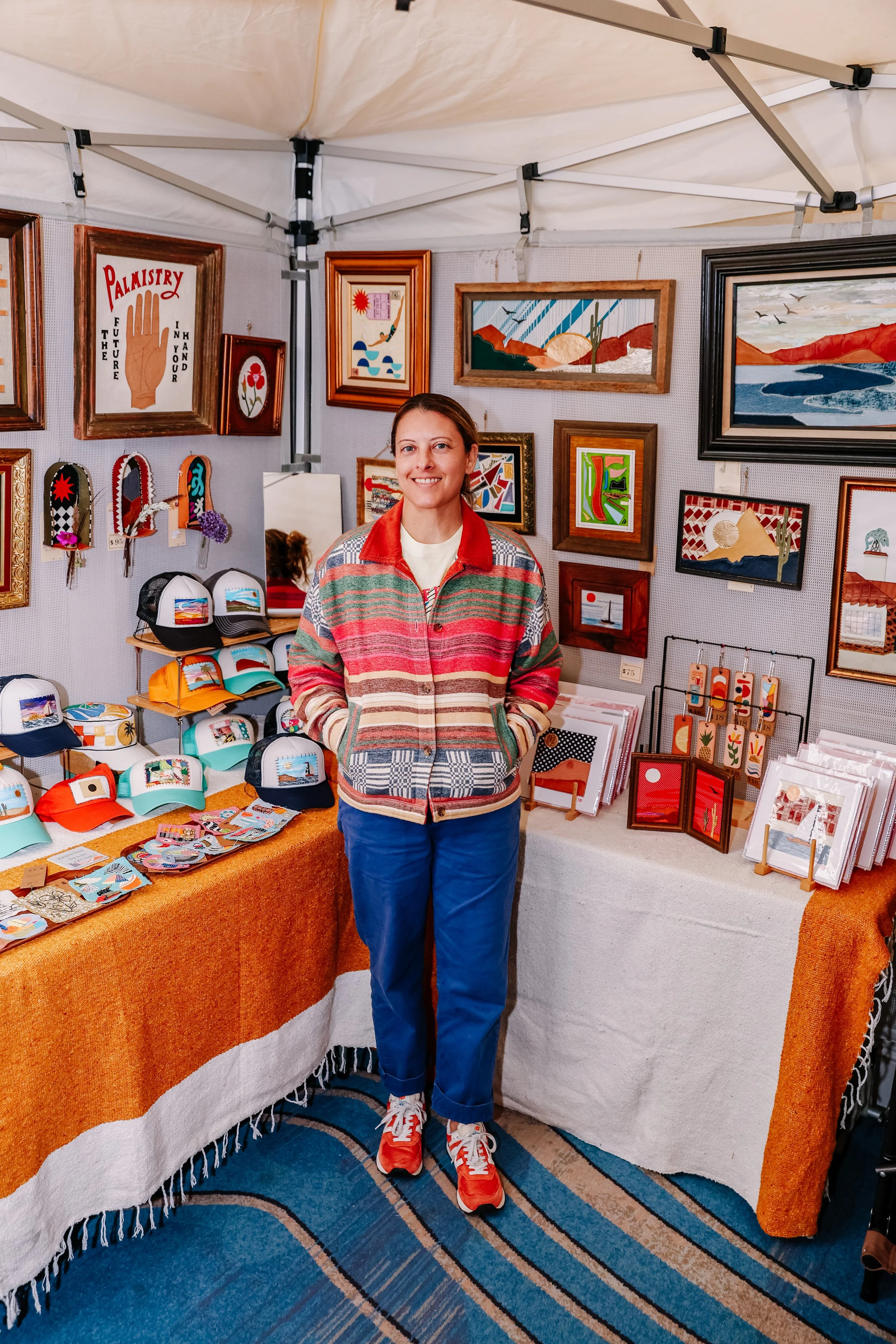 A woman standing at a booth displaying colorful handmade hats, jewelry, and framed artwork with desert and landscape scenes.