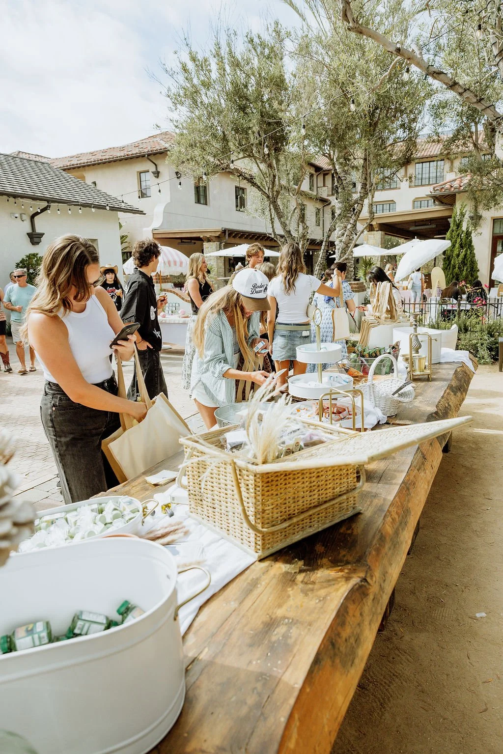 People shopping at an outdoor market table with various items, set in a sunny courtyard with trees and white house buildings in the background.