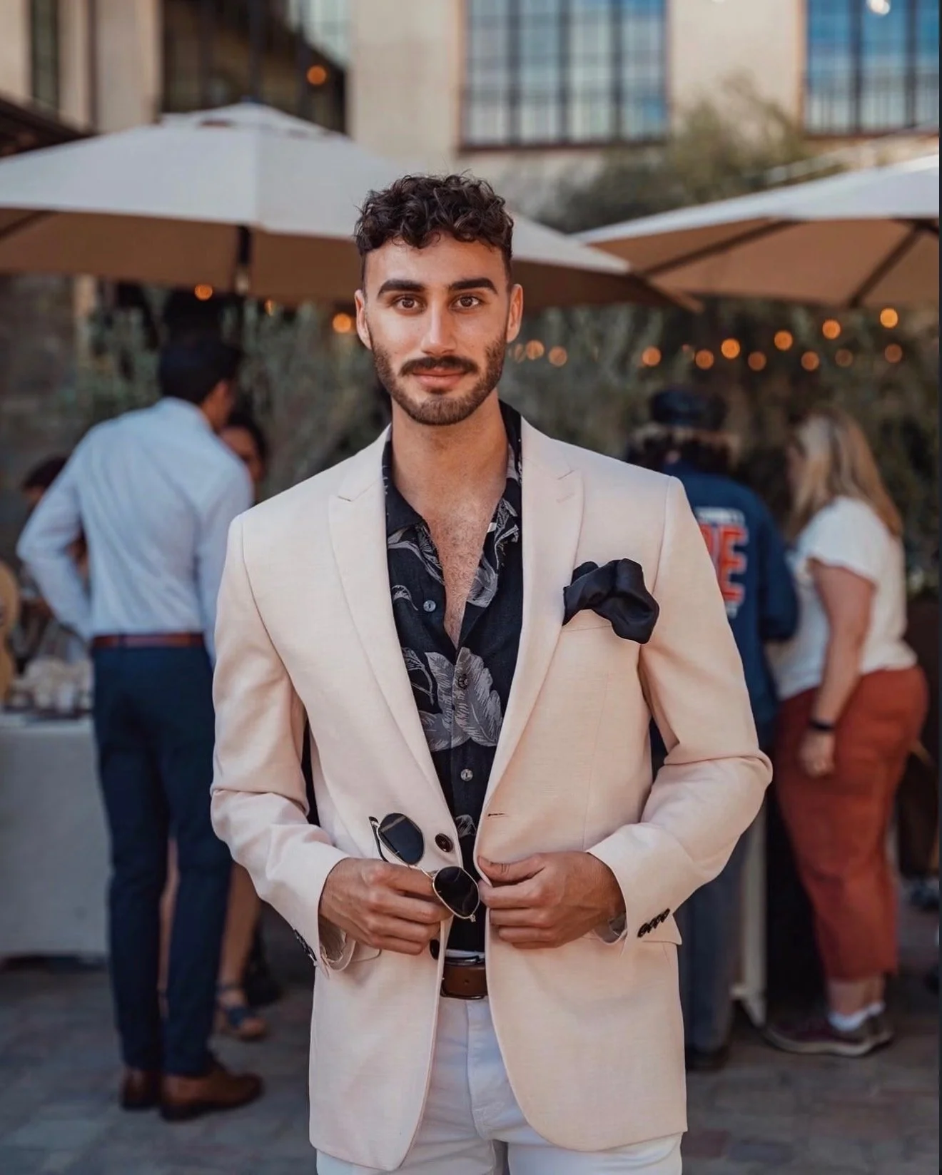 A young man with dark curly hair and a beard, dressed in a light-colored suit with a dark patterned shirt and a black pocket square, holding sunglasses, at an outdoor event with people and umbrellas in the background.