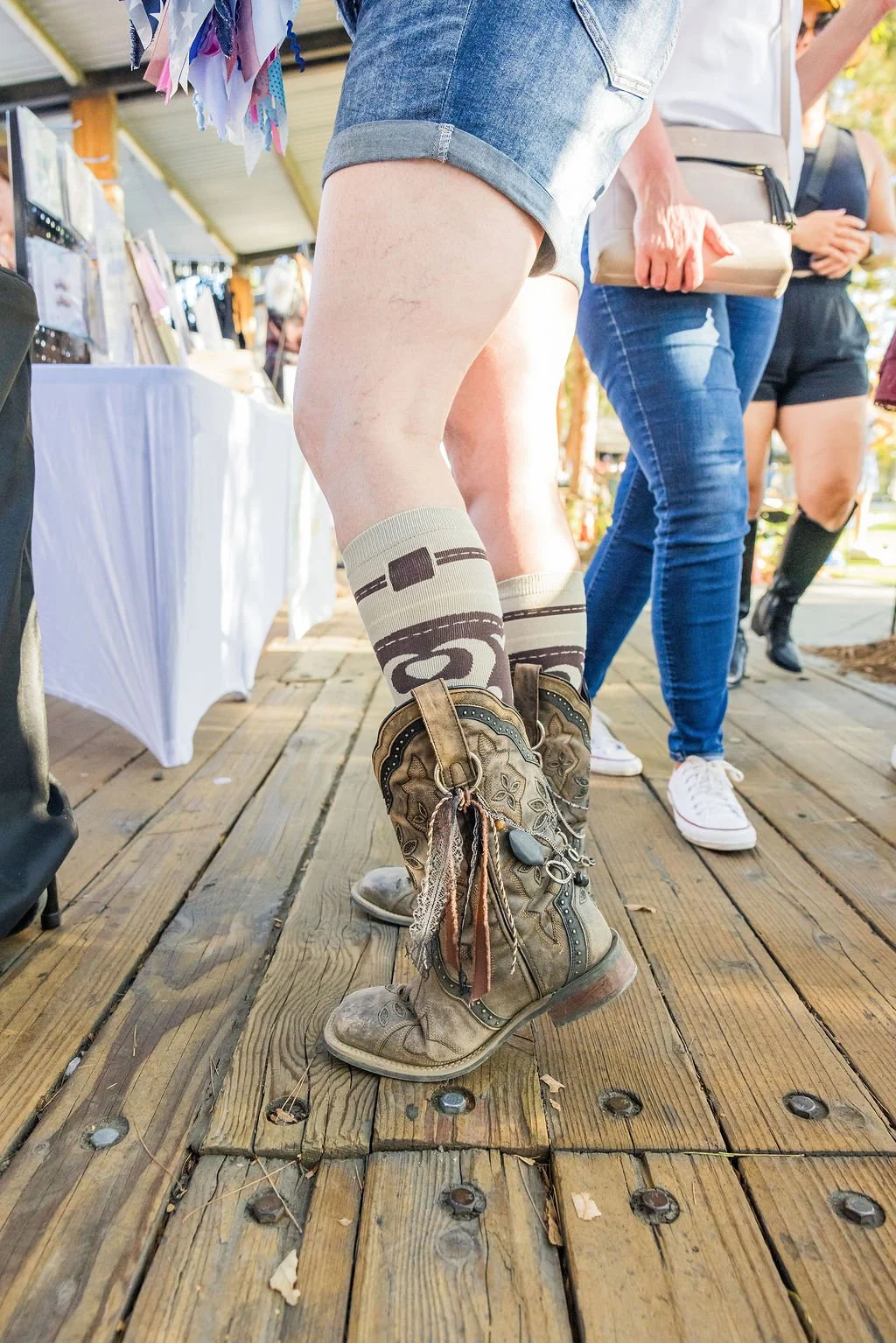 Close-up of a person standing on wooden porch wearing vintage cowboy boots and patterned knee-high socks, with other people and display tables visible in the background.