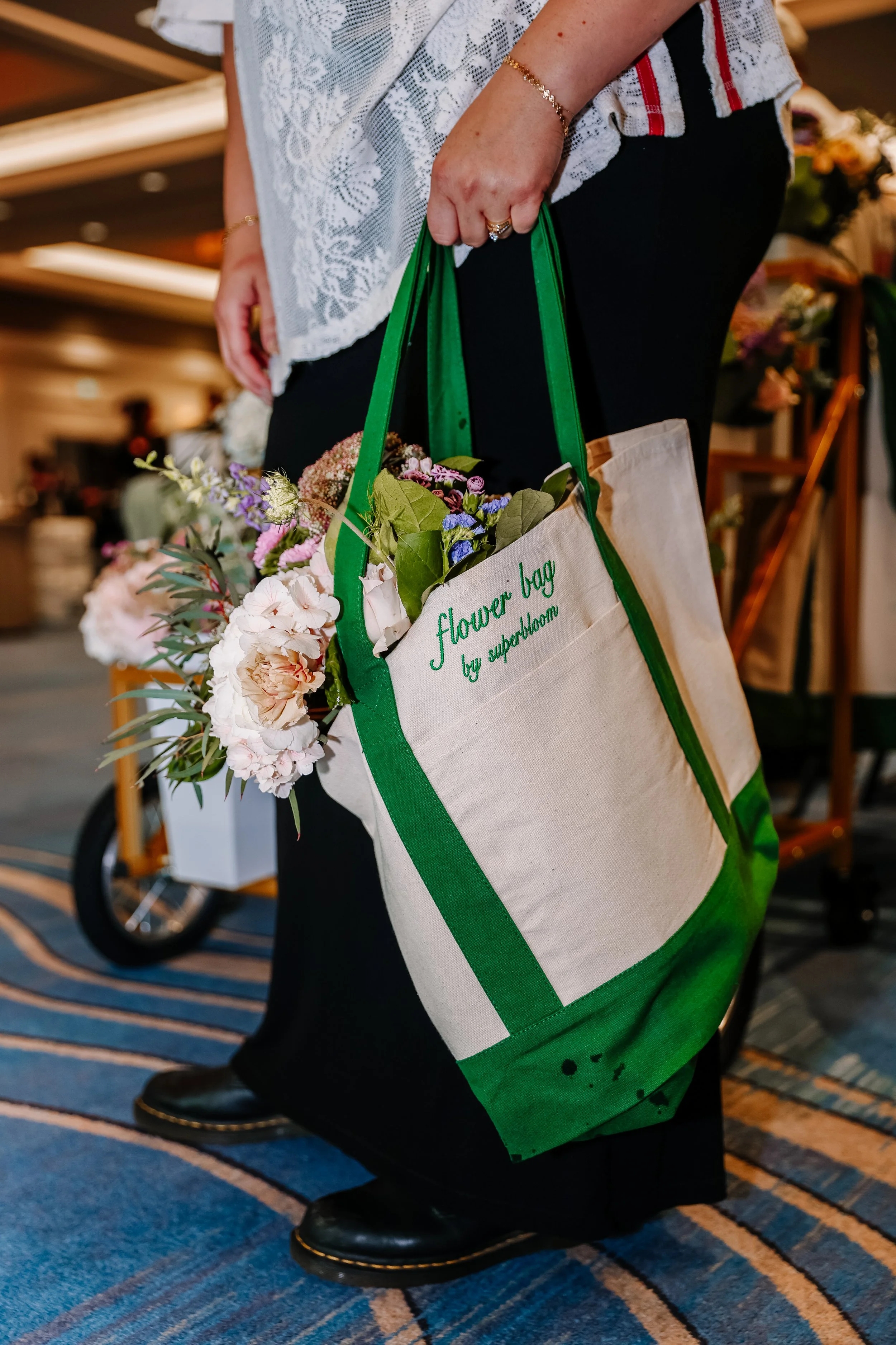 Person holding a beige cloth tote bag with green handles and green embroidered text that reads 'flower bag by superdawn,' filled with pink, purple, and white flowers. The person is wearing black pants, shiny black shoes with gold trim, a lacy white t