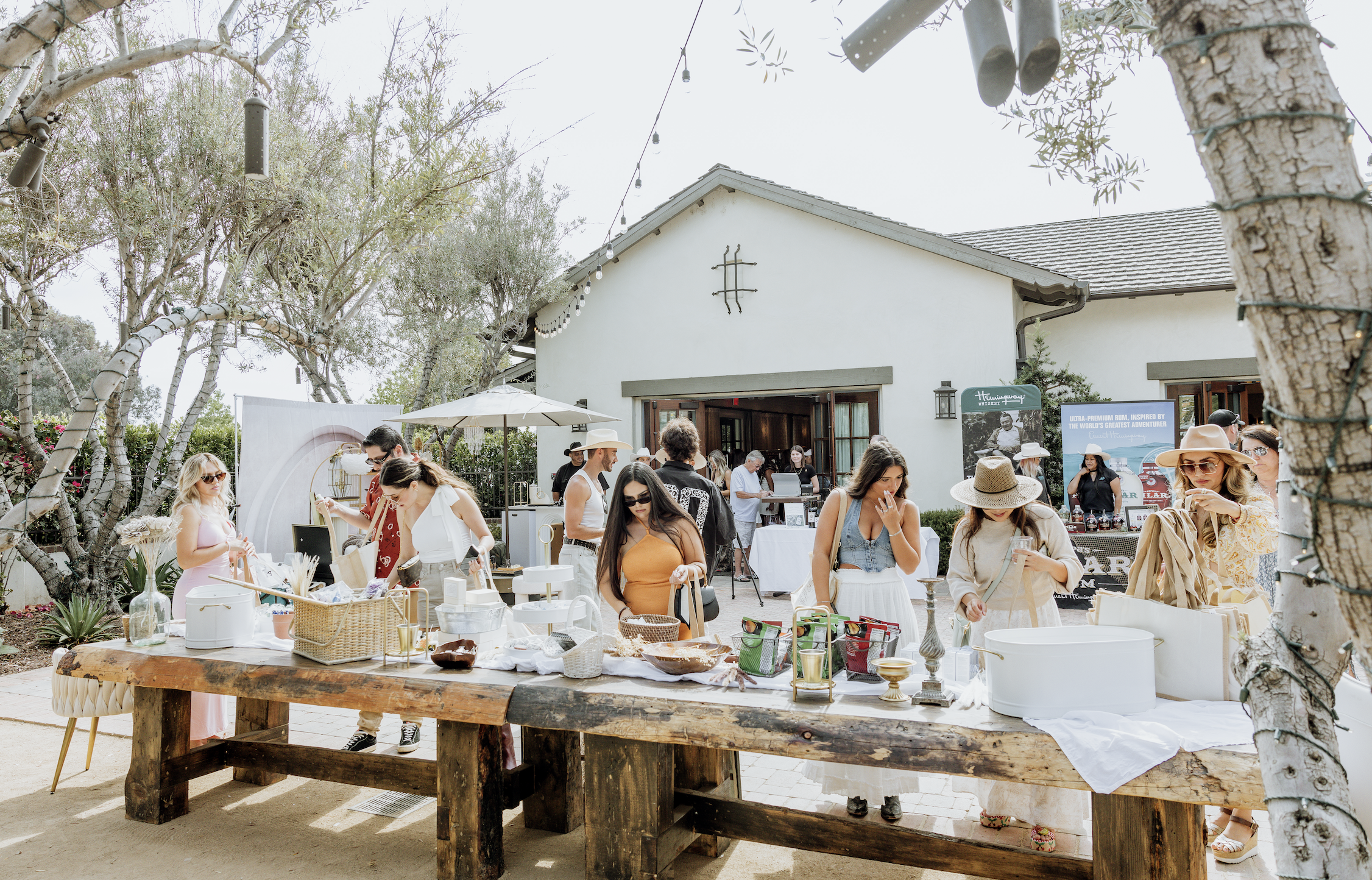 People browsing and shopping at a market or outdoor event in a sunny backyard setting with trees and a white building in the background.