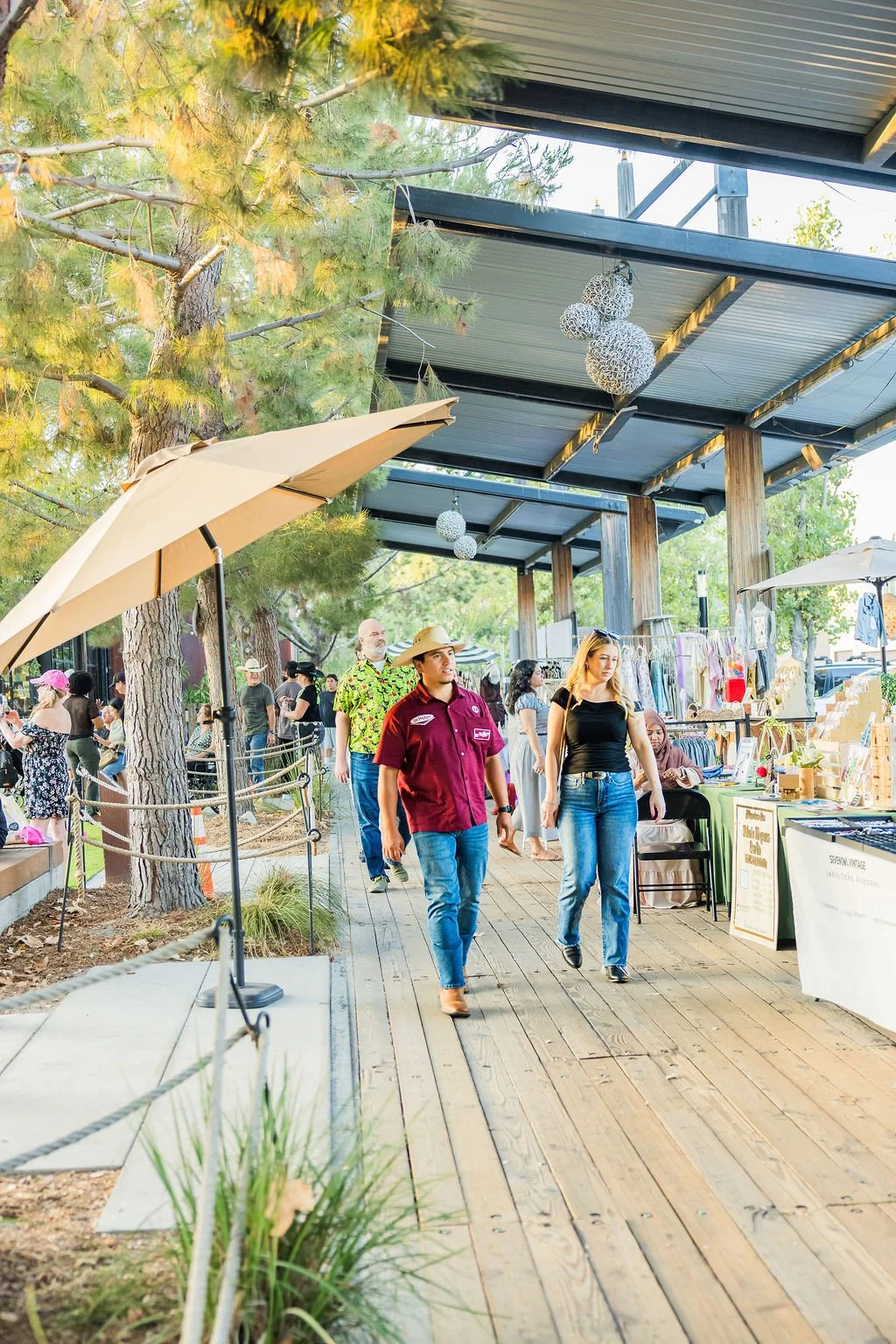 People walking along an outdoor shopping walkway with stalls, trees, umbrellas, and hanging decorative lights.