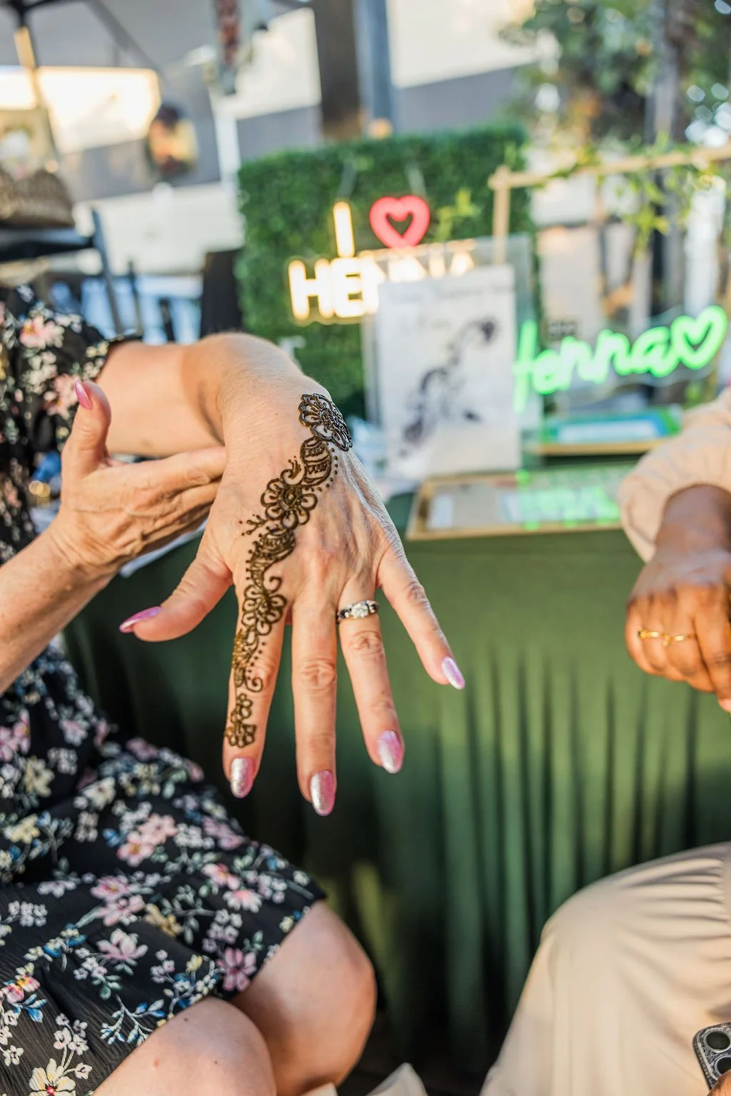 Two women showing off their decorated hands, one with henna design and wearing rings, in a setting with neon signs that say 'I love HEAVEN' and 'HENNA' in the background.