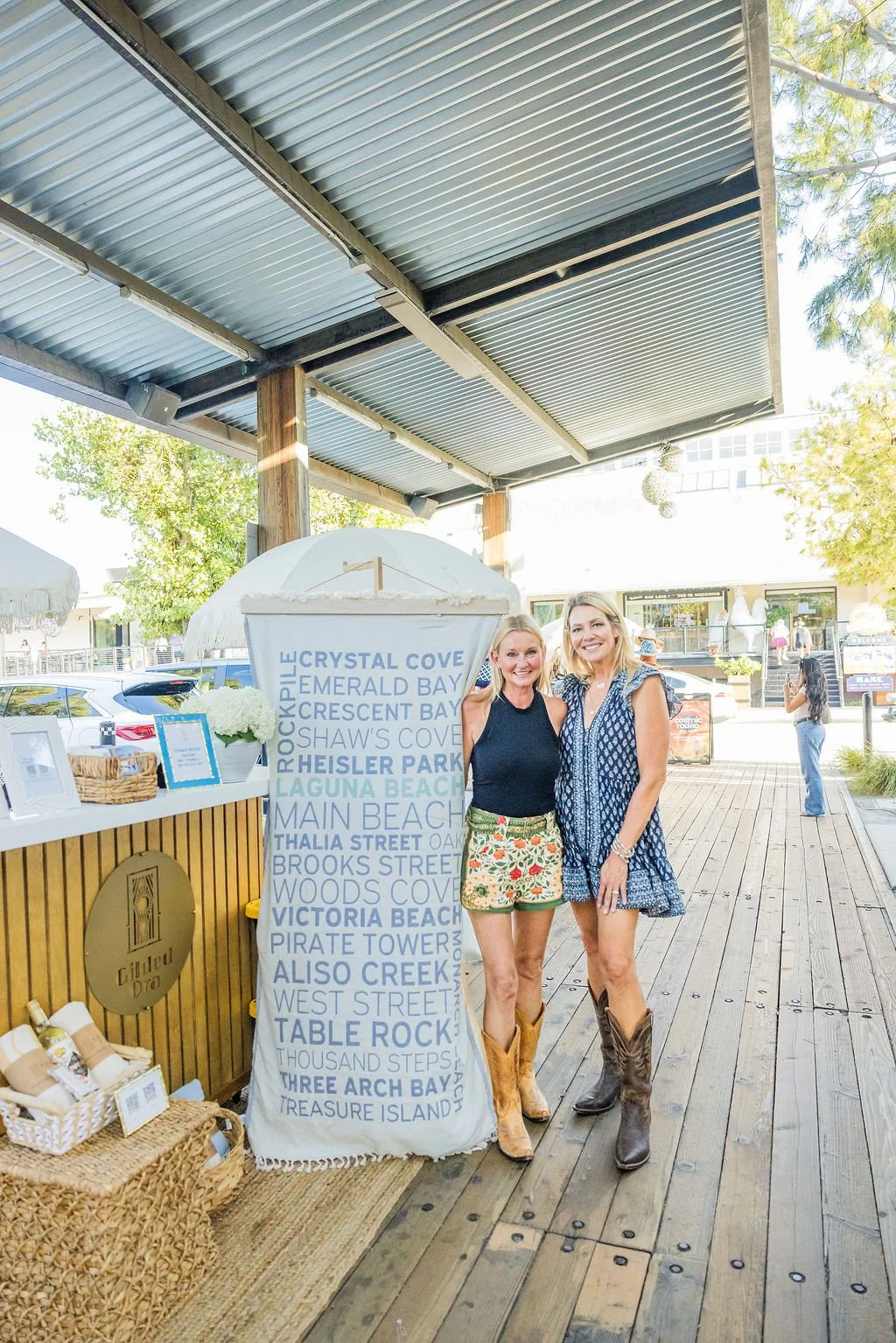 Two women standing outdoors near a sign with local beach and street names, smiling at the camera. One woman is wearing a navy sleeveless top and floral shorts, the other is in a blue patterned dress. There is a wooden booth and some cars in the backg