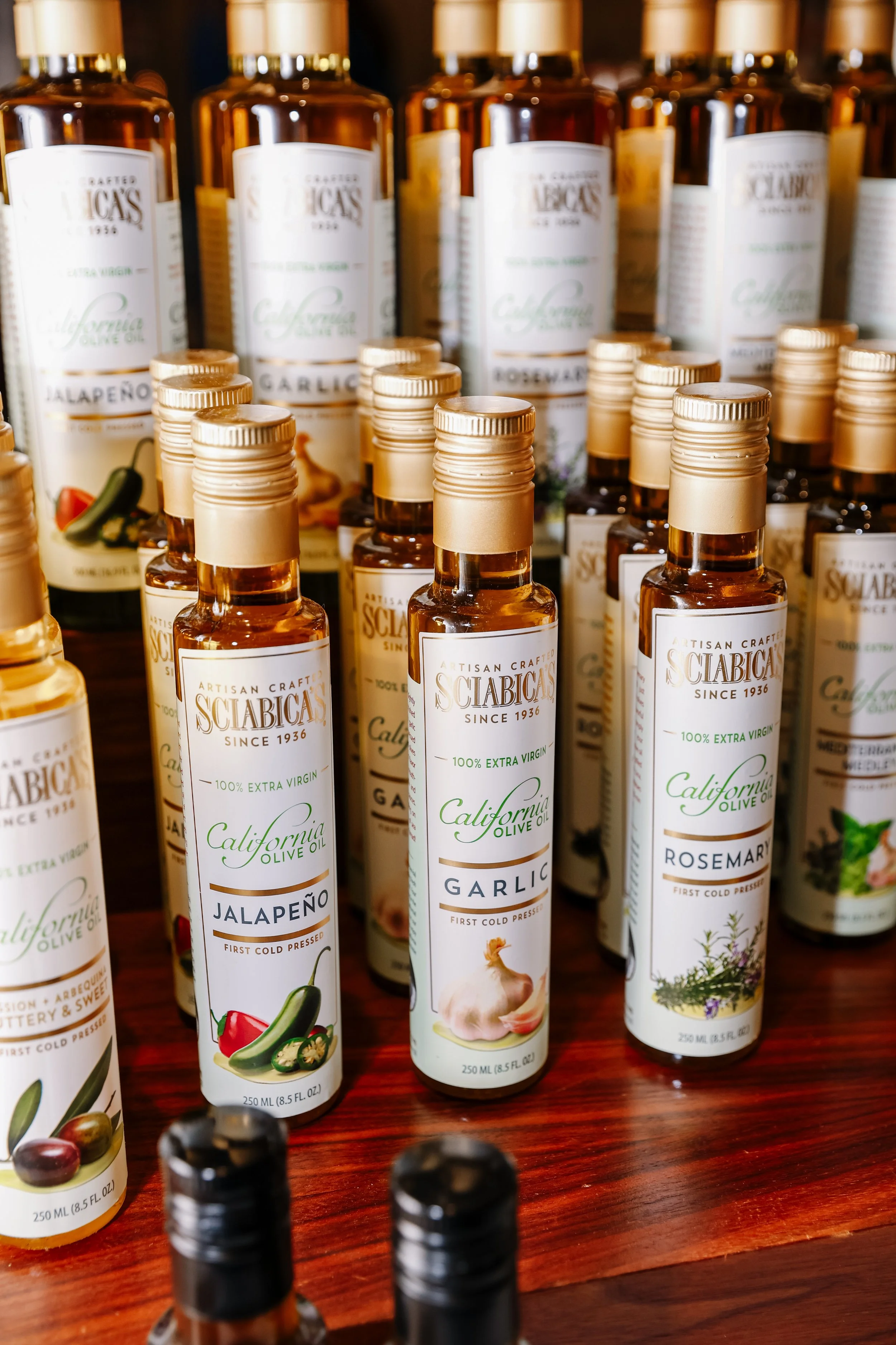 Display of bottles of artisan crafted California olive oil with flavors like garlic, jalapeño, rosemary, and garlic, arranged on a wooden surface.