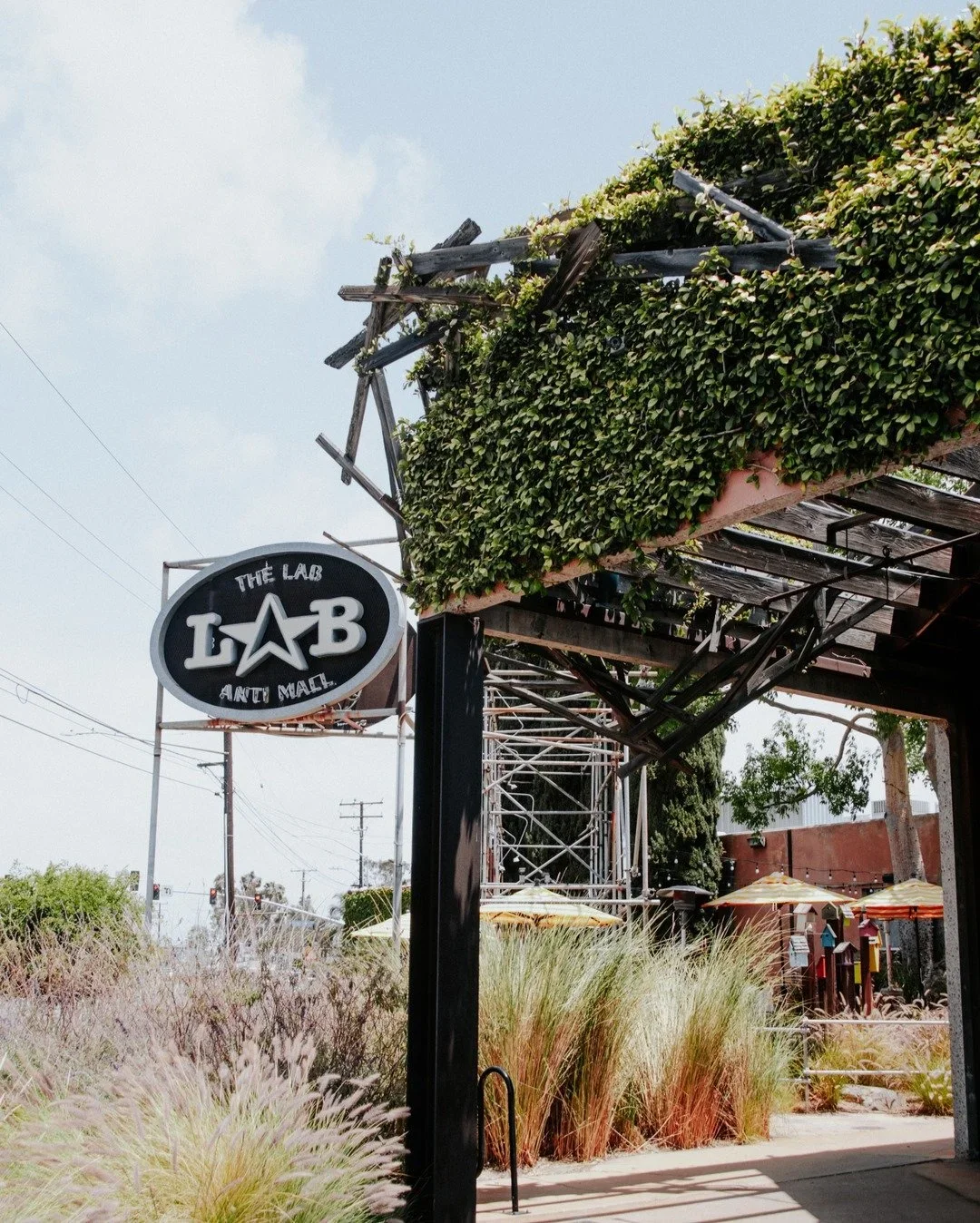 A sign for The Lab Anti Mall with an overgrown roof, surrounded by tall grasses and outdoor umbrellas.