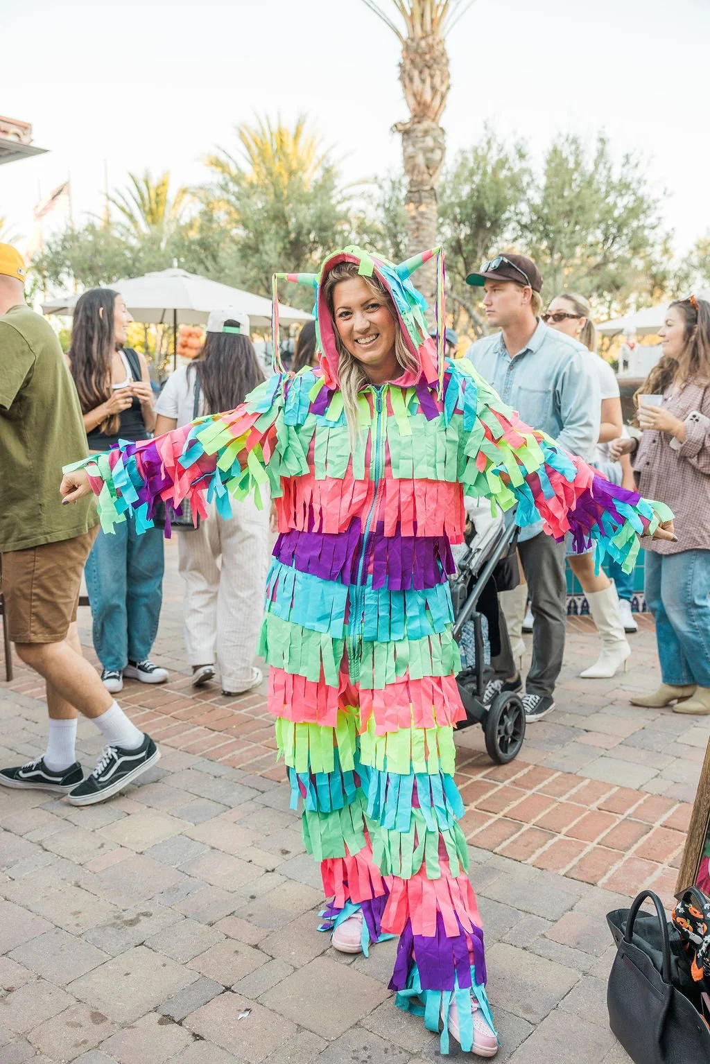 Woman smiling in a colorful, fringed llama costume at an outdoor event with people in the background.