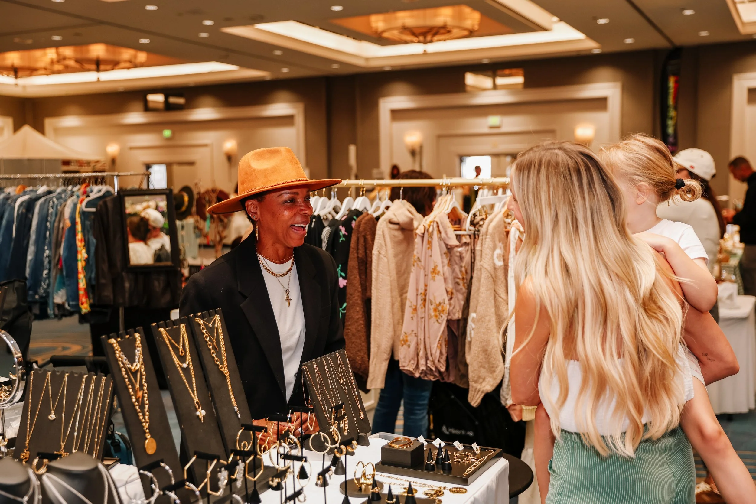 A vendor with a brown hat and gold jewelry displays jewelry at a booth while a young woman with long blonde hair and a white tank top looks at the jewelry at an indoor market.