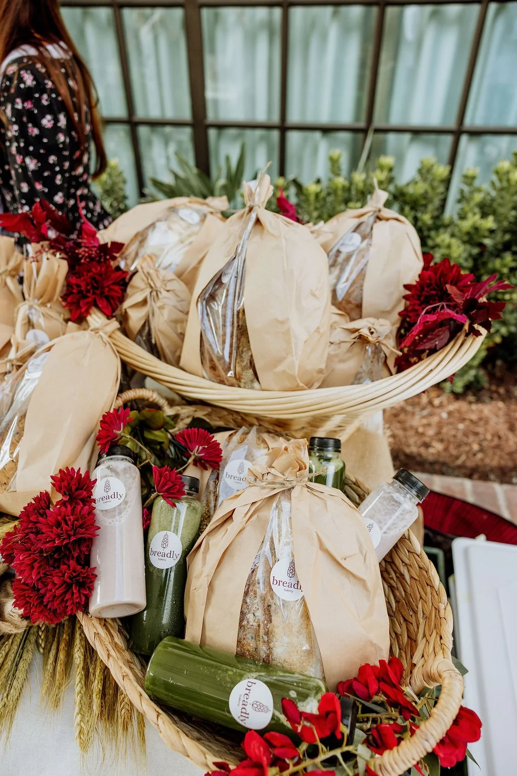 Basket filled with wrapped loaves of bread, some with clear windows, and bottles of liquid, accompanied by red flowers and green foliage, displayed on a table.