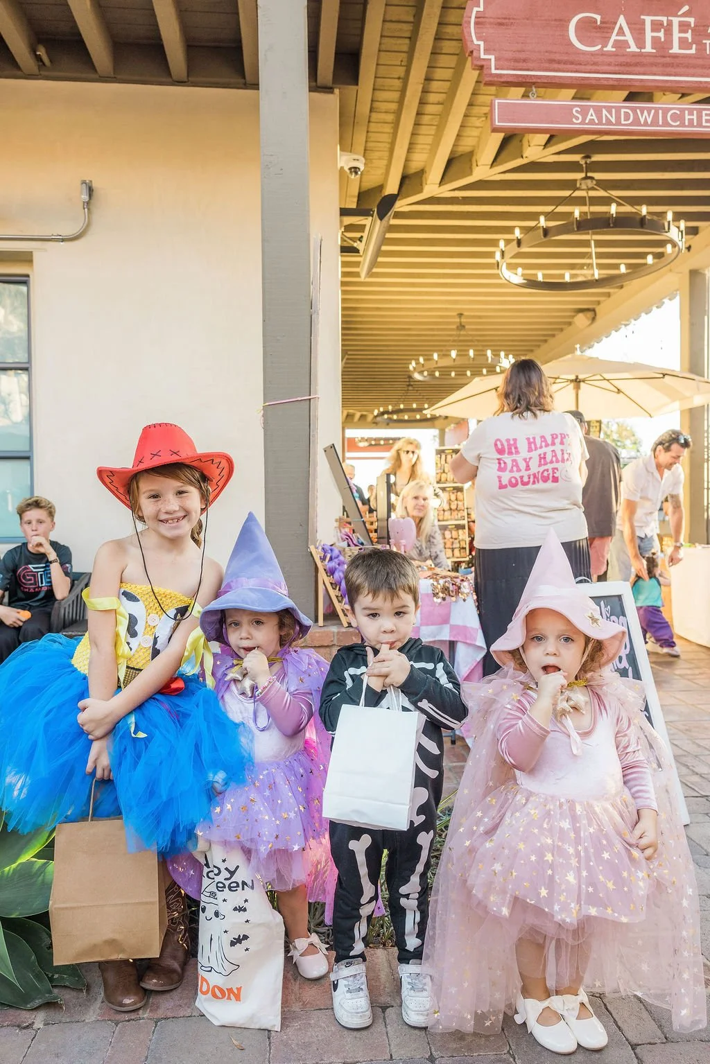 Four children dressed in Halloween costumes standing outdoors at a daytime event, with two boys and two girls, holding shopping bags and candy.