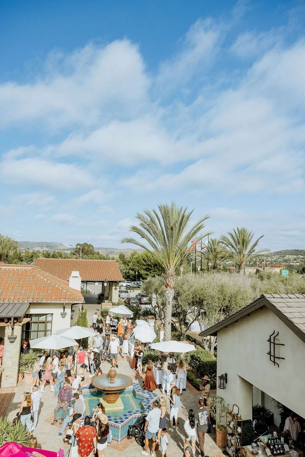 Outdoor gathering at a courtyard with people, palm trees, umbrellas, a fountain, and buildings with tiled roofs under a partly cloudy sky.