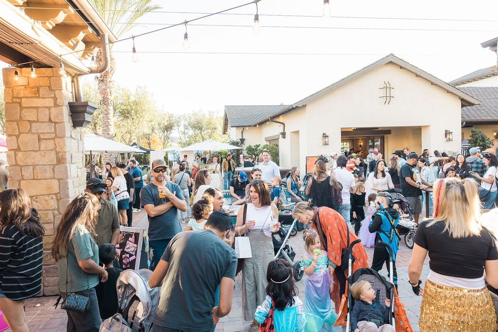 Crowd of families and children outside a building during the daytime, some dressed as princesses and superheroes, with children in strollers, enjoying an outdoor event with string lights overhead.