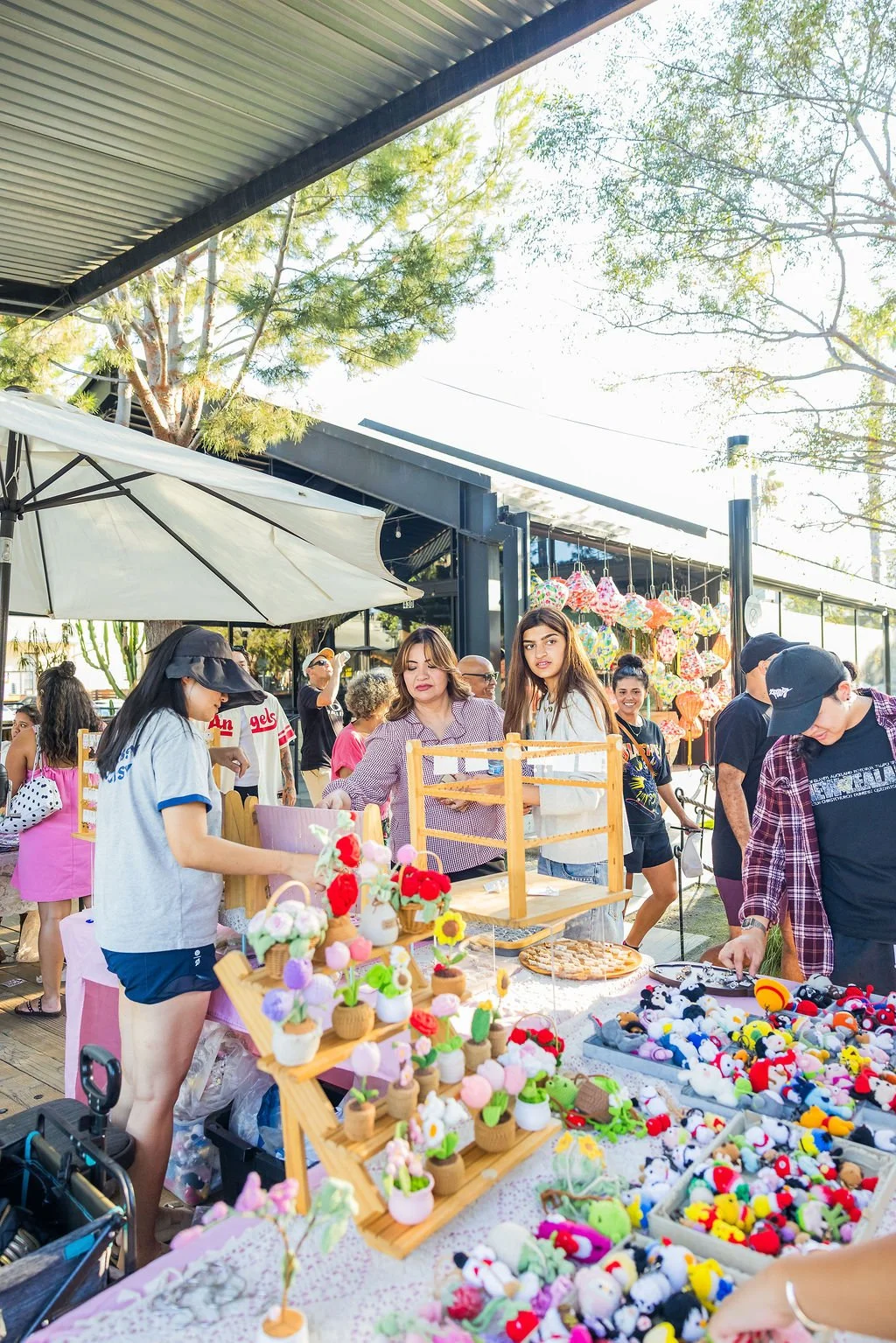People shopping at an outdoor market stall with colorful handmade crafts, including felt animals and decorative items, under a covered area with trees and colorful lanterns in the background.
