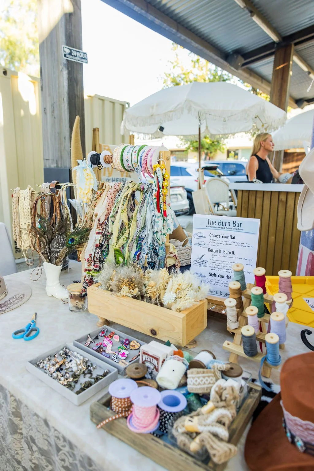 A market stall selling accessories such as hairbands, scarves, jewelry, and sewing supplies under umbrellas. A woman stands behind the stall, and a sign provides instructions for customizing hats.