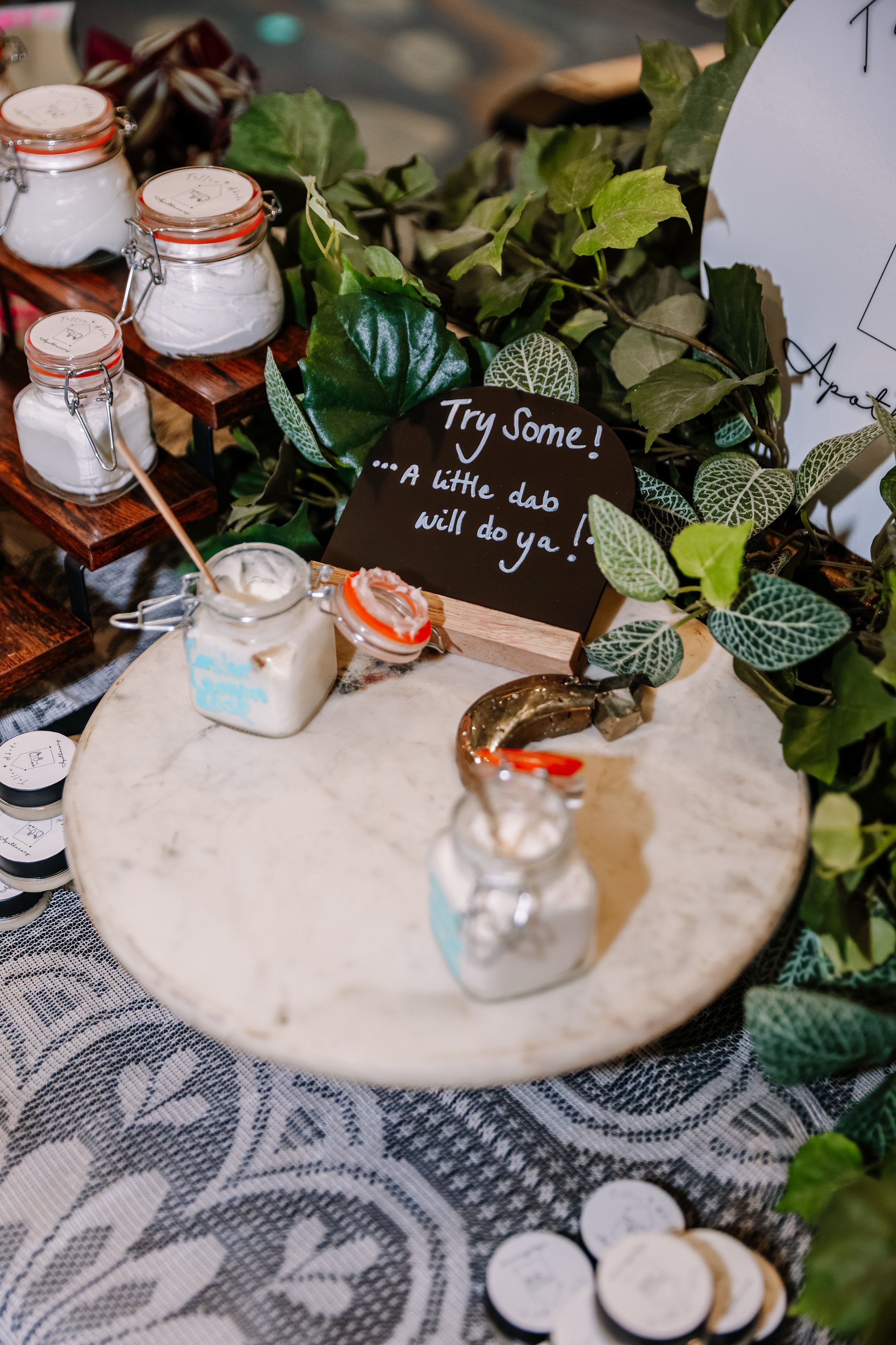 Small glass jars filled with white powdered substance, some with orange rubber seals, placed on a lace tablecloth surrounded by green foliage and a decorative brass animal figure. A sign reads 'Try Some! A Little dab will do ya!'
