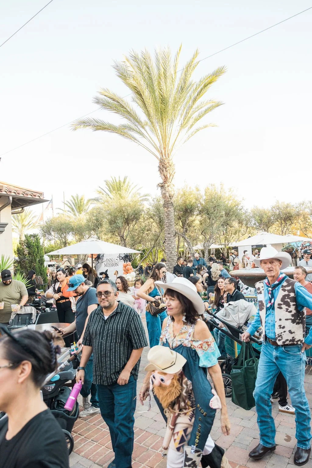 Crowd of people gathered outdoors under a tall palm tree, some pushing strollers, others socializing, with umbrellas and trees in the background.