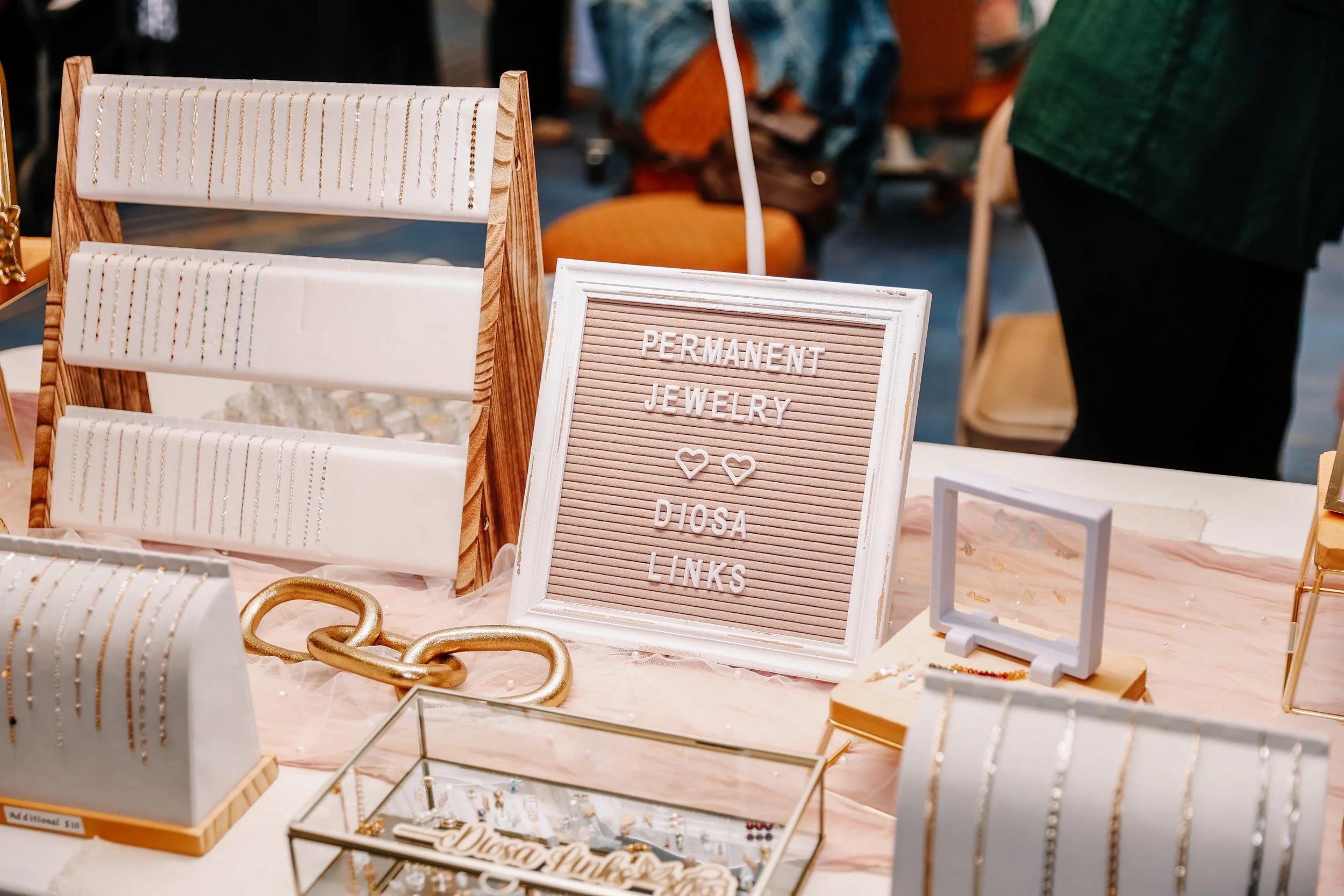 Jewelry display at a craft fair with a sign that reads 'Permanent Jewelry Diosa Links' and various jewelry pieces including necklaces and bracelets.