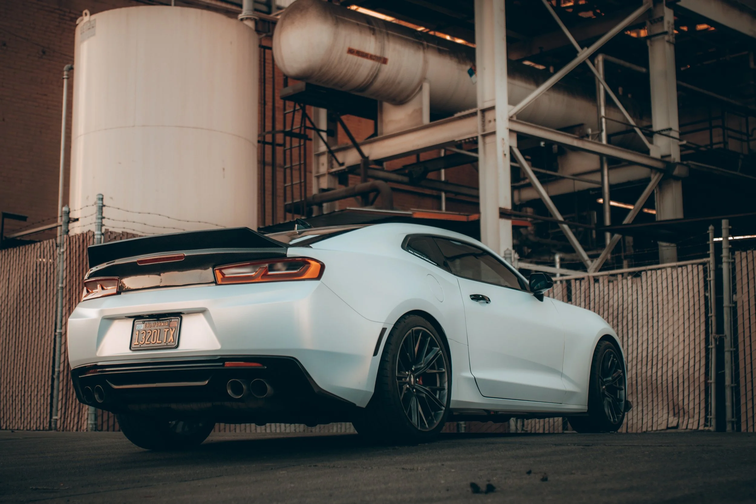 A white sports car with a black rear spoiler and black wheels parked in front of an industrial building with large tanks and metal pipes.