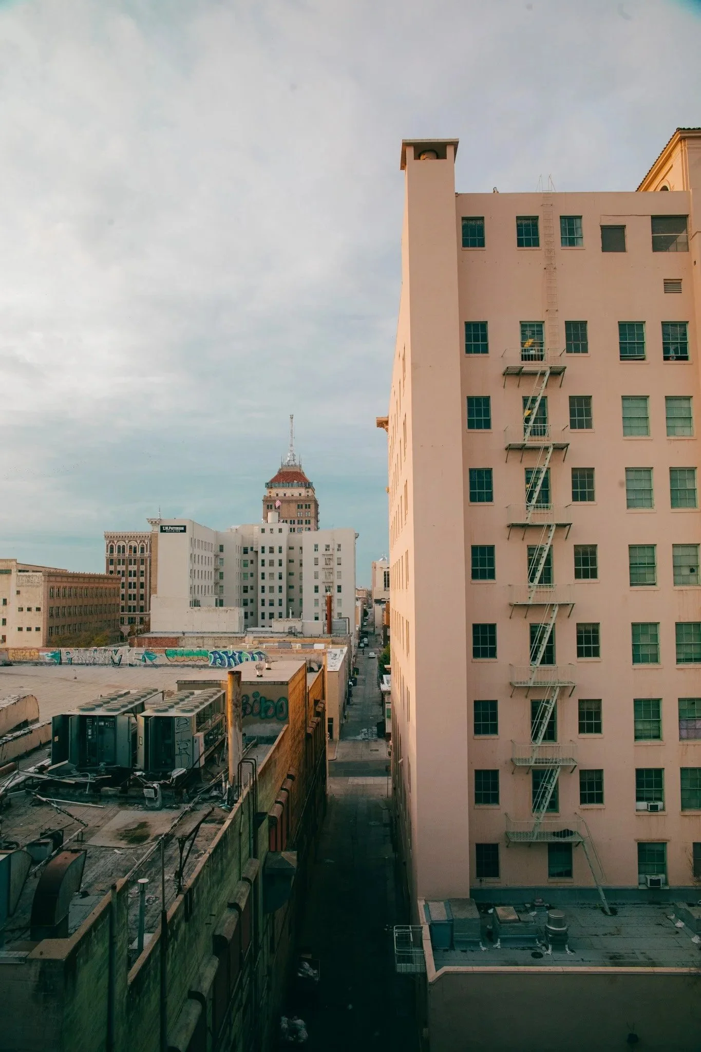 View of high-rise buildings in an urban cityscape during daytime. The image shows a tall, peach-colored building with a fire escape on the right side, and other buildings with city billboards and graffiti on rooftops background. The sky is cloudy.
