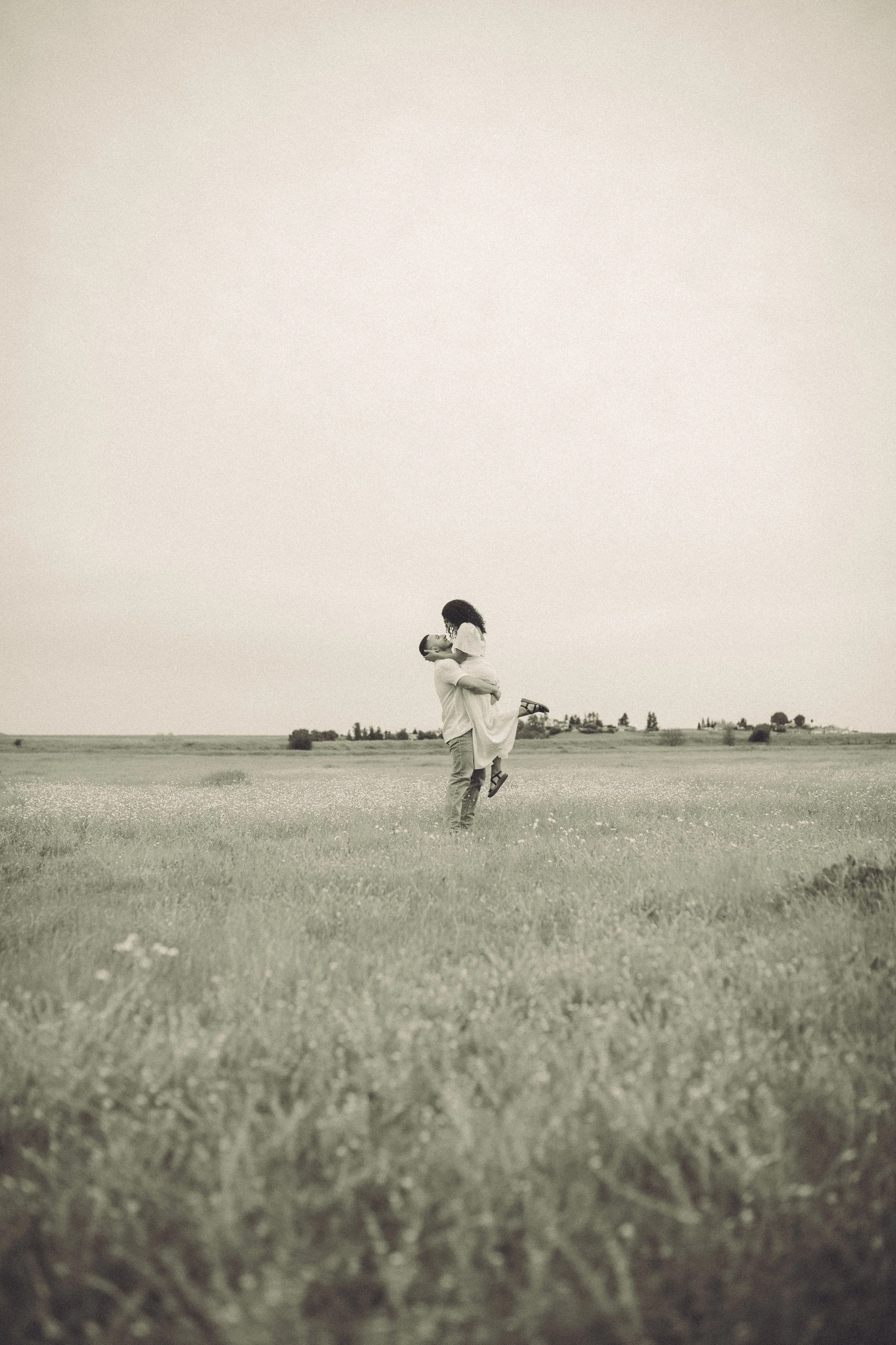 A couple is standing in a large open field, with the man lifting the woman in his arms. The woman is wearing a light-colored dress and sandals, and the man is dressed casually. The sky is clear and the trees are visible in the distant background.