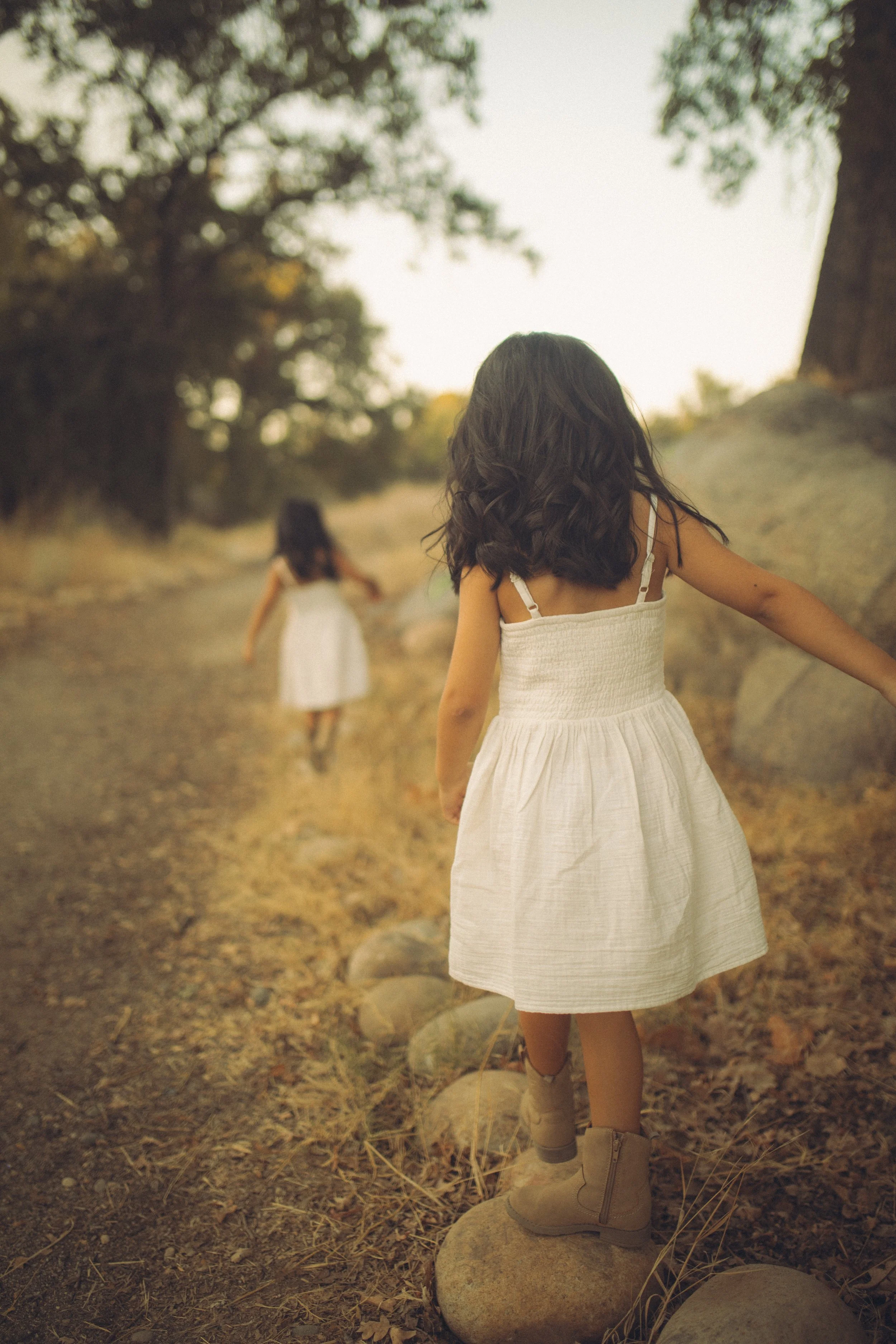 Two girls wearing white dresses walking outdoors on a rocky path surrounded by trees in warm sunlight.