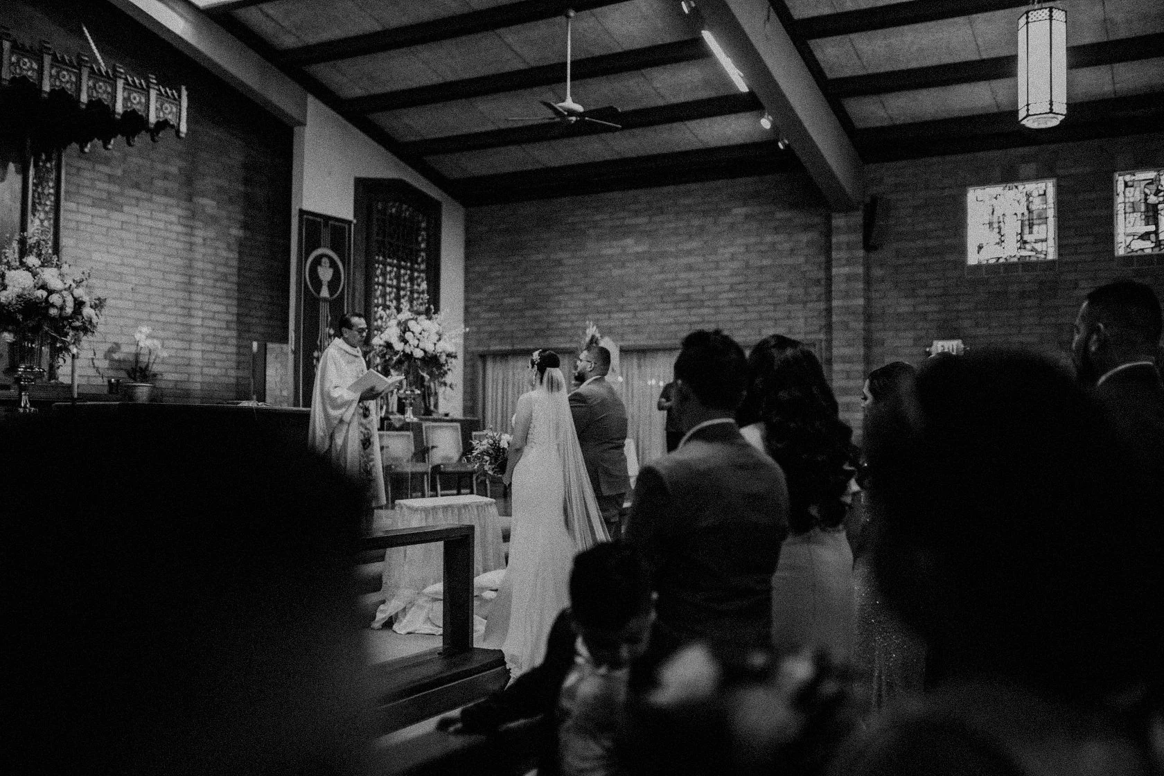 A black and white photo of a wedding ceremony inside a church. The bride and groom stand facing a priest, who is reading from a book. Guests sit in the foreground, watching the ceremony. There are flowers and religious symbols on the altar in the bac