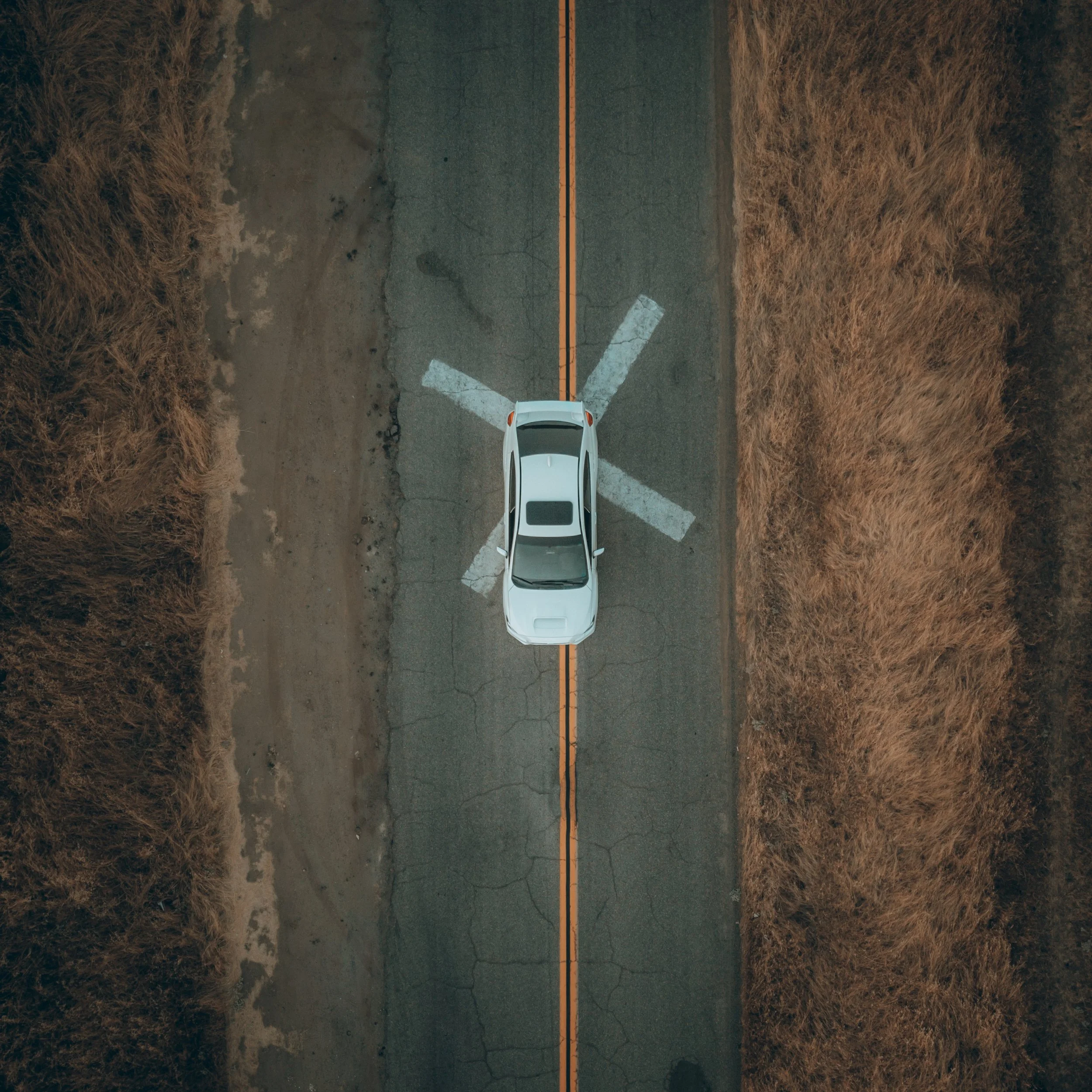 Top-down view of a white car on a cracked asphalt road with a yellow double line in the center, surrounded by dry, brown grass.