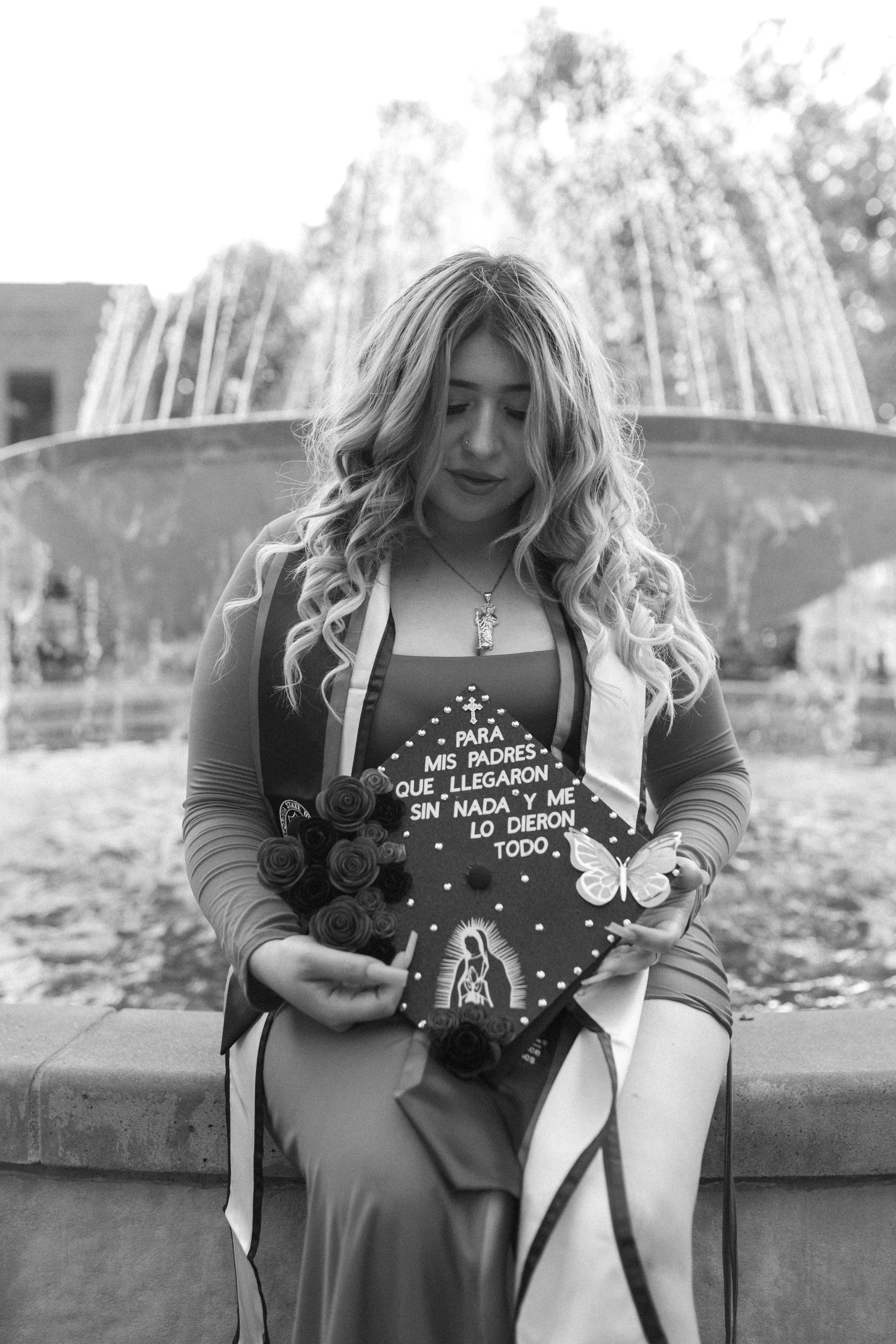 A woman with long curly hair, wearing a dress and jewelry, holding a sign with a religious image, a bouquet of roses, and a butterfly decoration outdoors in front of a fountain.