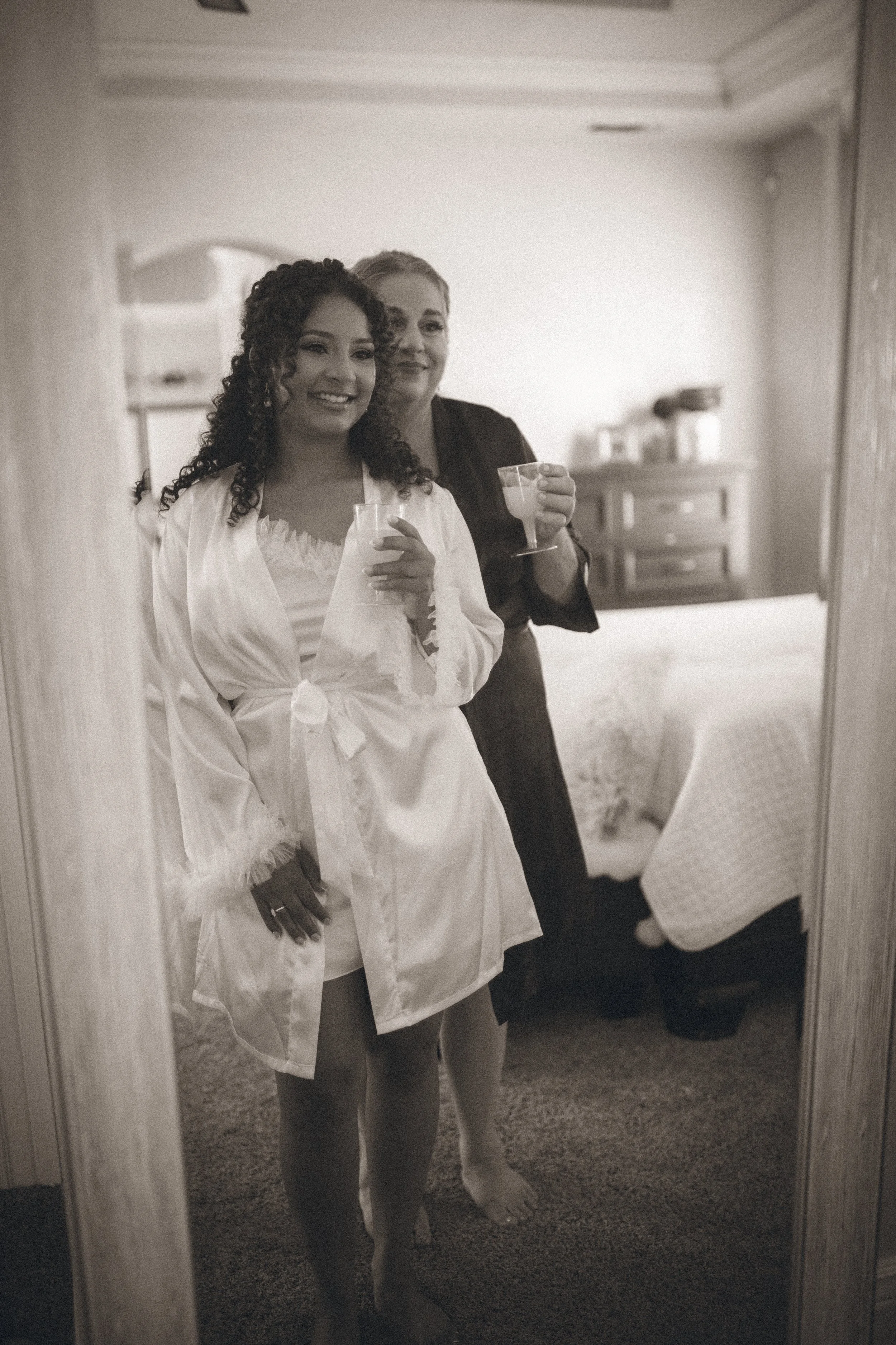 A woman in white satin robe with lace trim and a woman in black top with drinks in hand, smiling, taking a mirror selfie in a bedroom.