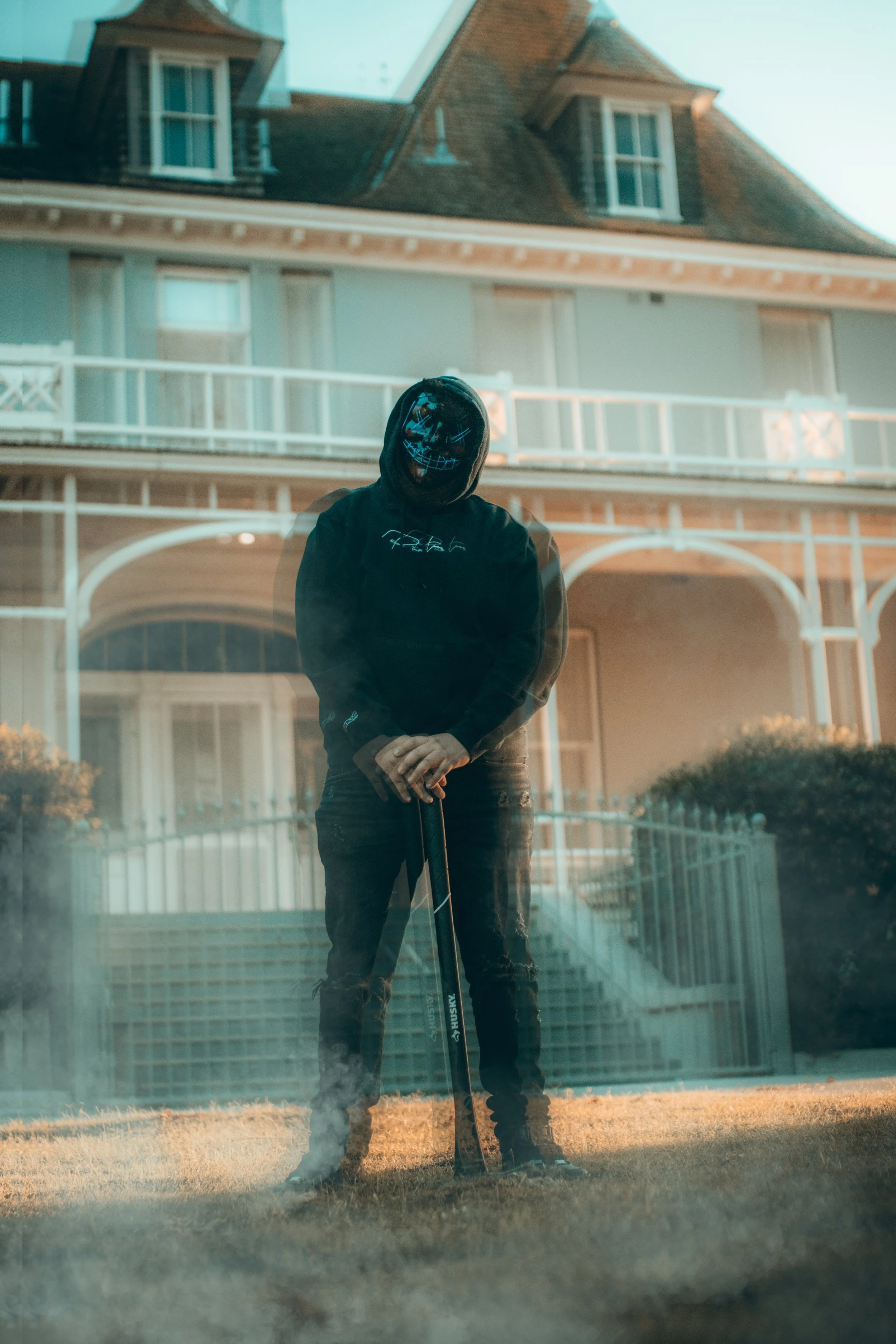 Reflection of a person wearing a black hoodie with a glowing skull mask, standing outside on the grass, holding a golf club, in front of a house with a balcony and dormer windows.