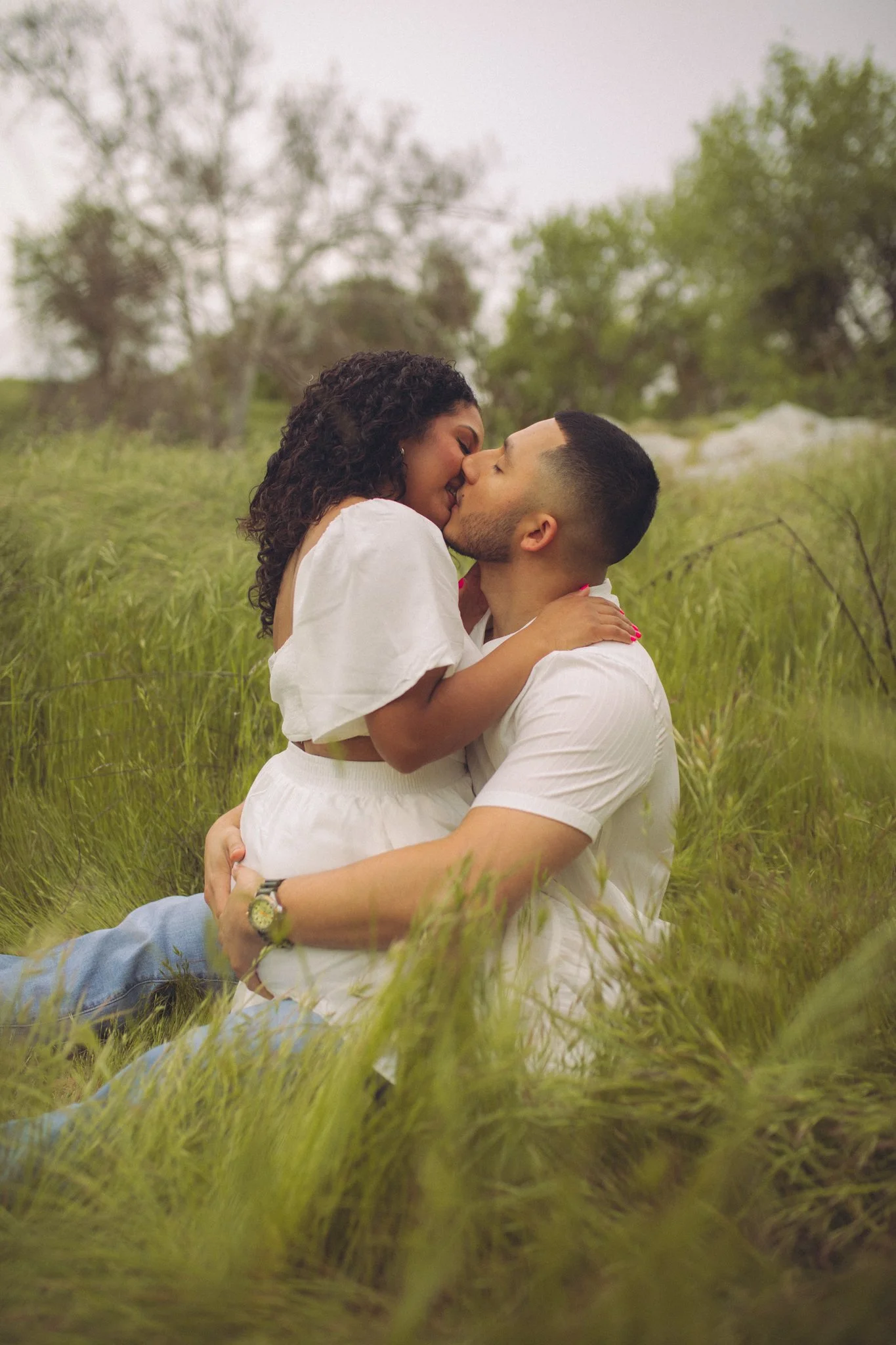 A man and woman in a grassy field, kissing and hugging each other, with trees in the background.