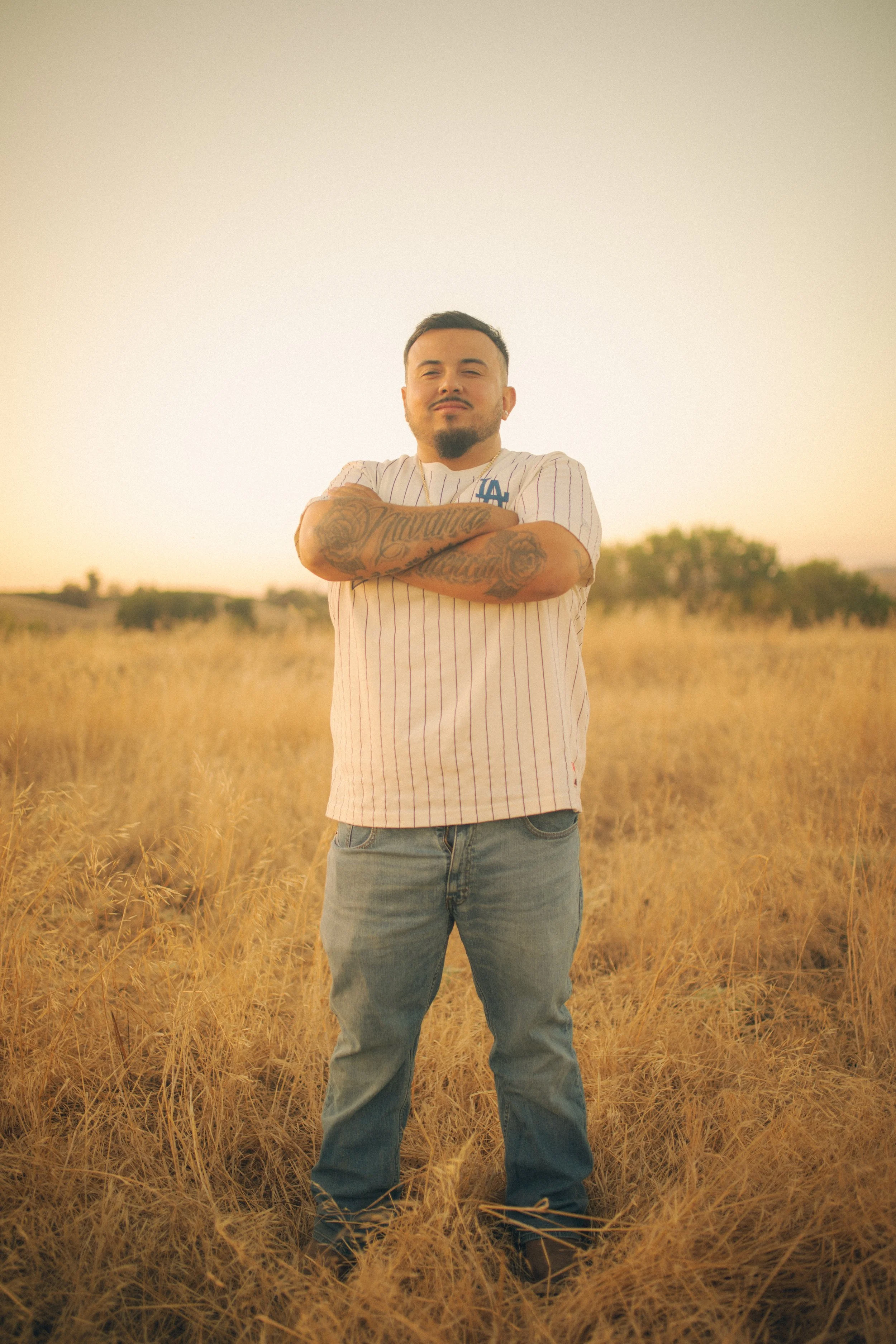 A man with tattoos on his arms and a goatee standing in a field of tall grass at sunset, arms crossed, looking at the camera.