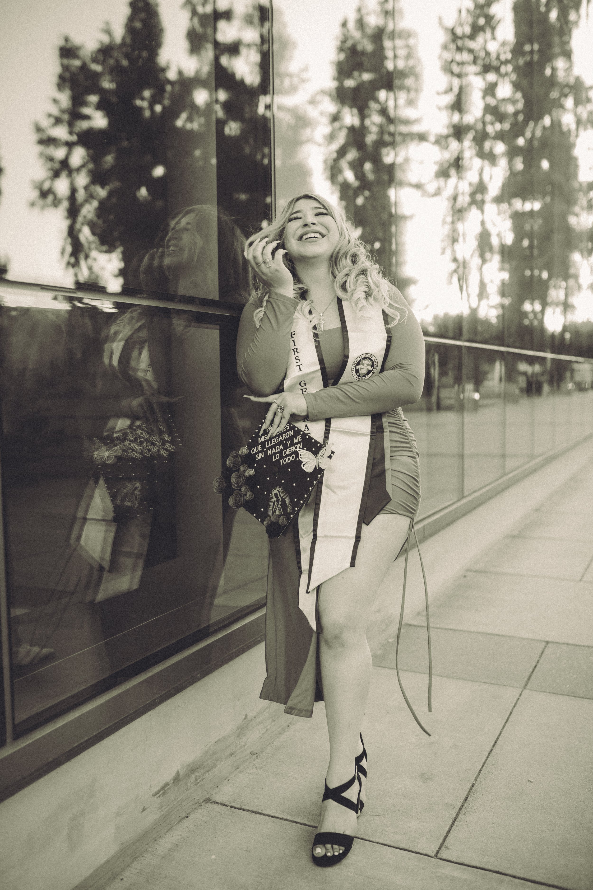 A woman with curly hair and a sash that reads 'First Generation' is smiling and laughing with her head tilted back, leaning against a reflective glass building, outdoors during the daytime.