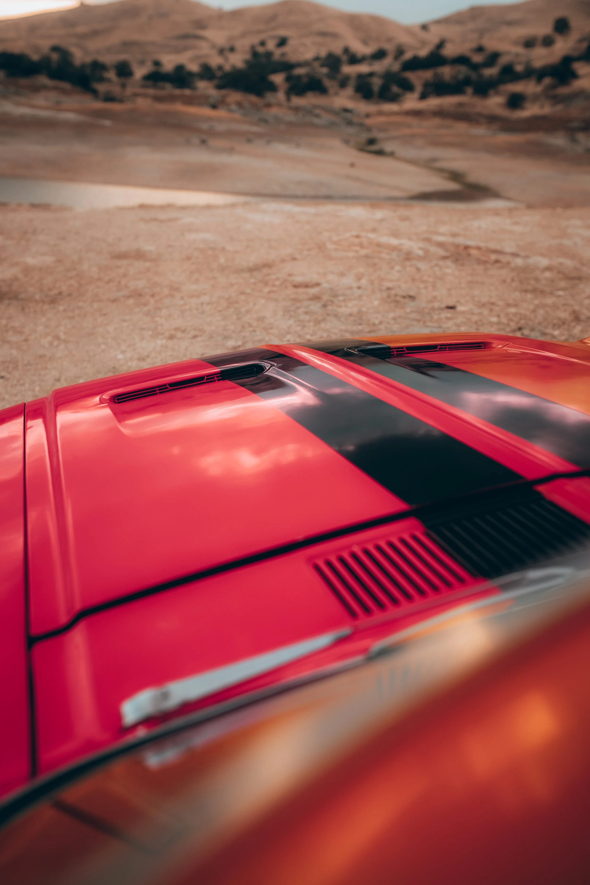 The image shows the front part of a red vehicle with black racing stripes on the hood, with a desert landscape in the background featuring sandy terrain and rolling hills.