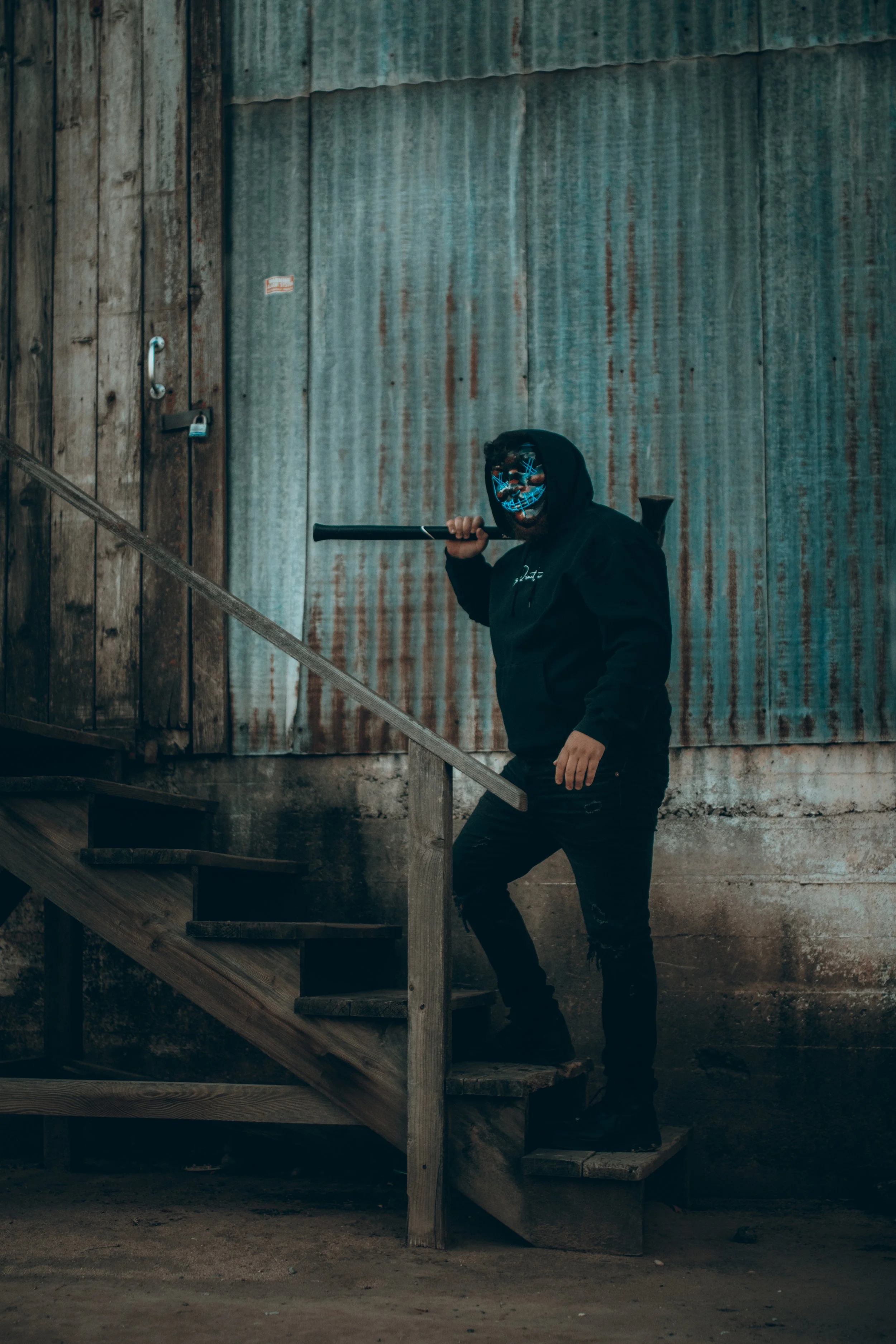 A person wearing a black hoodie and dark pants, holding a bat, standing on a wooden staircase in front of a rustic metal wall. The person is wearing a mask with neon blue and orange design.