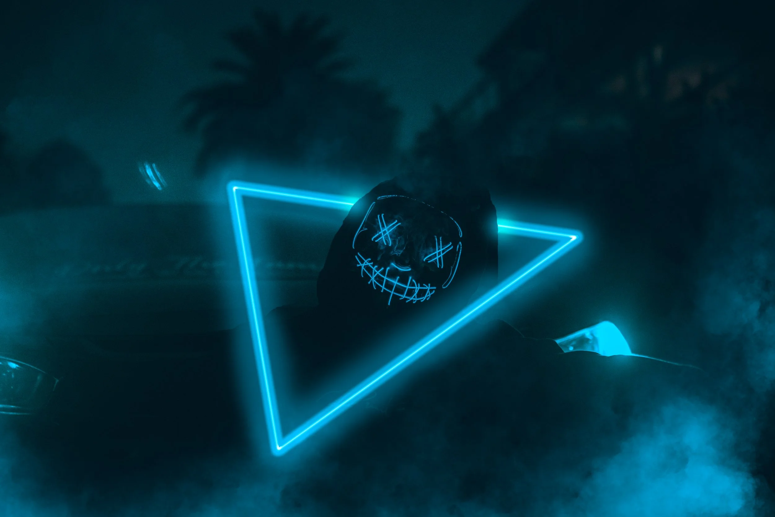 A person wearing a glowing, illuminated mask with a smiling skull face, illuminated blue triangle, and neon blue lights in a dark environment.