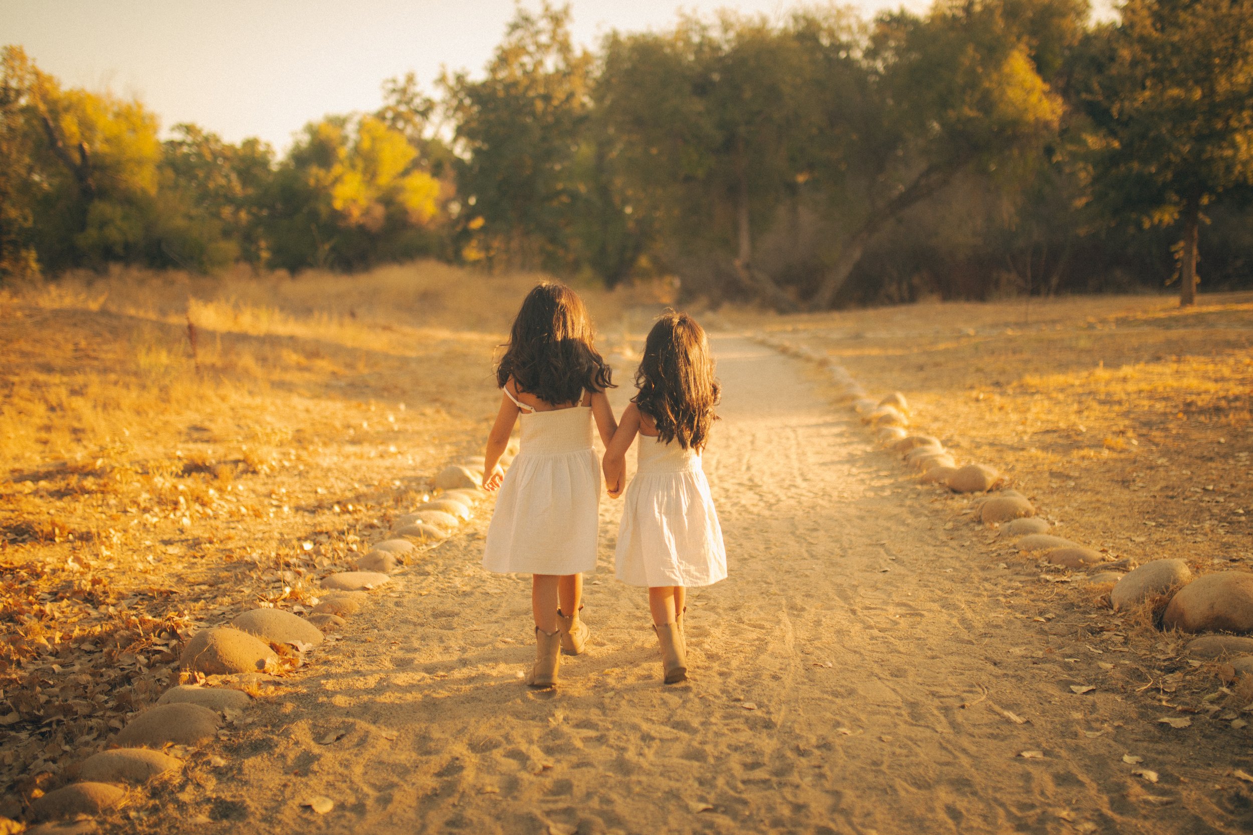 Two young girls in white dresses walking hand in hand down a dirt path surrounded by trees during autumn.
