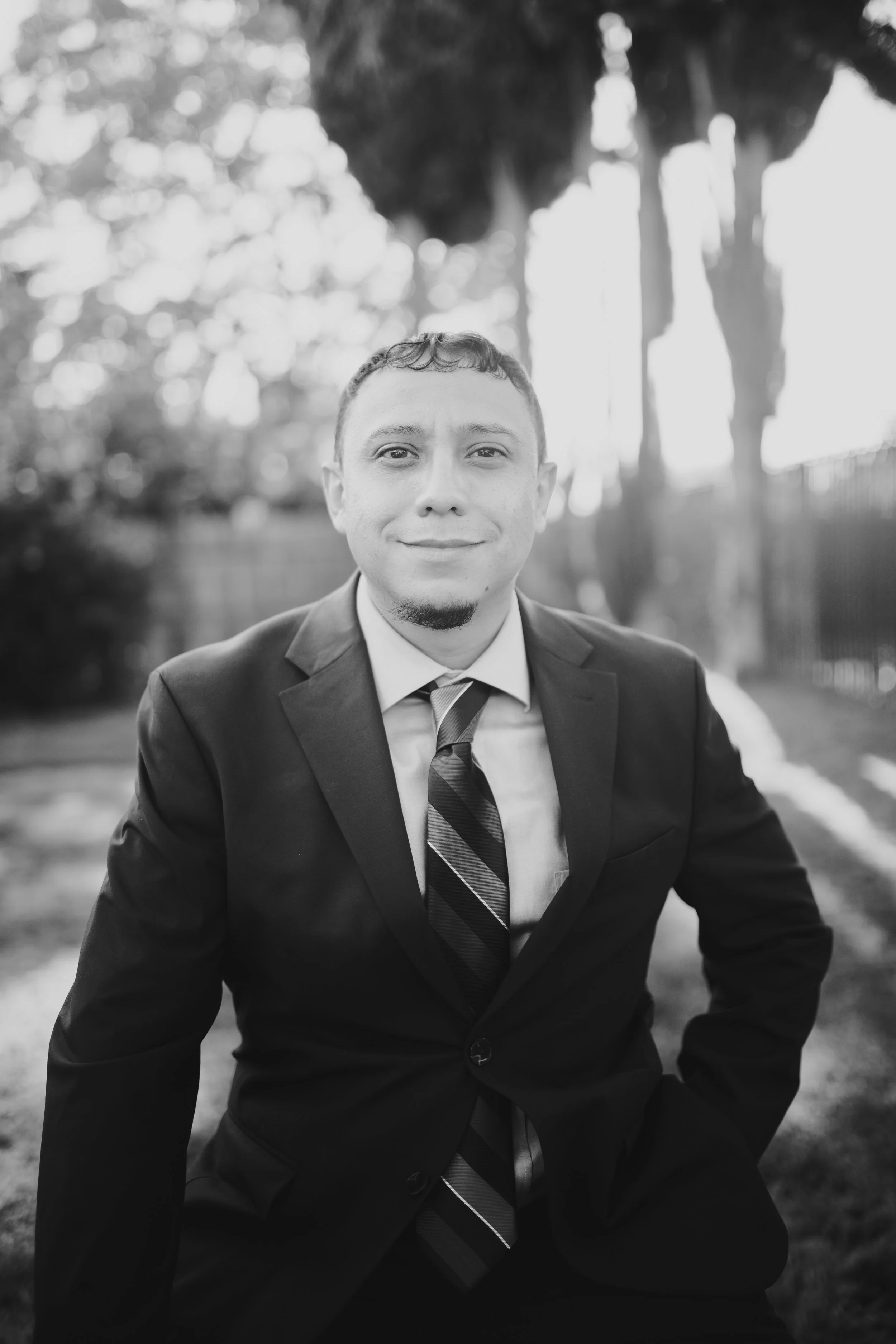Black and white portrait of a man in a suit and tie, smiling outdoors with trees in the background.