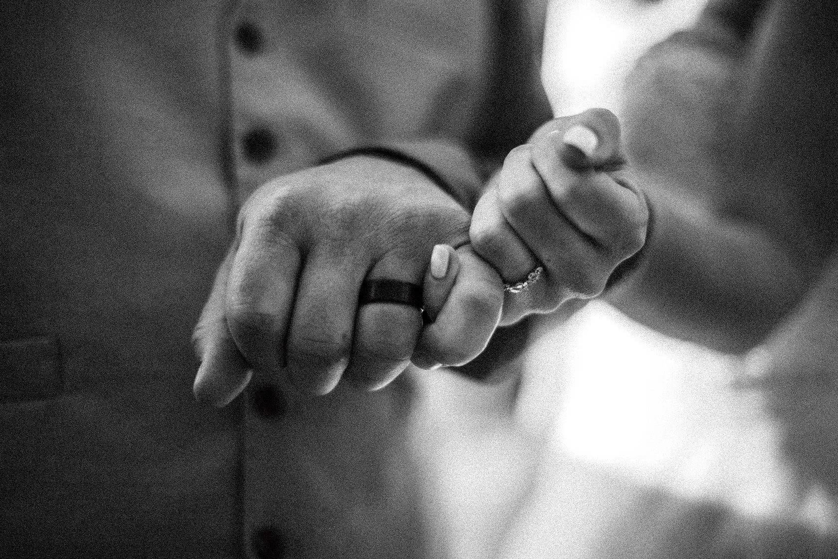 Close-up of two hands grasping each other, with one hand wearing a black ring on the middle finger and the other hand wearing a ring on the ring finger, in black and white.