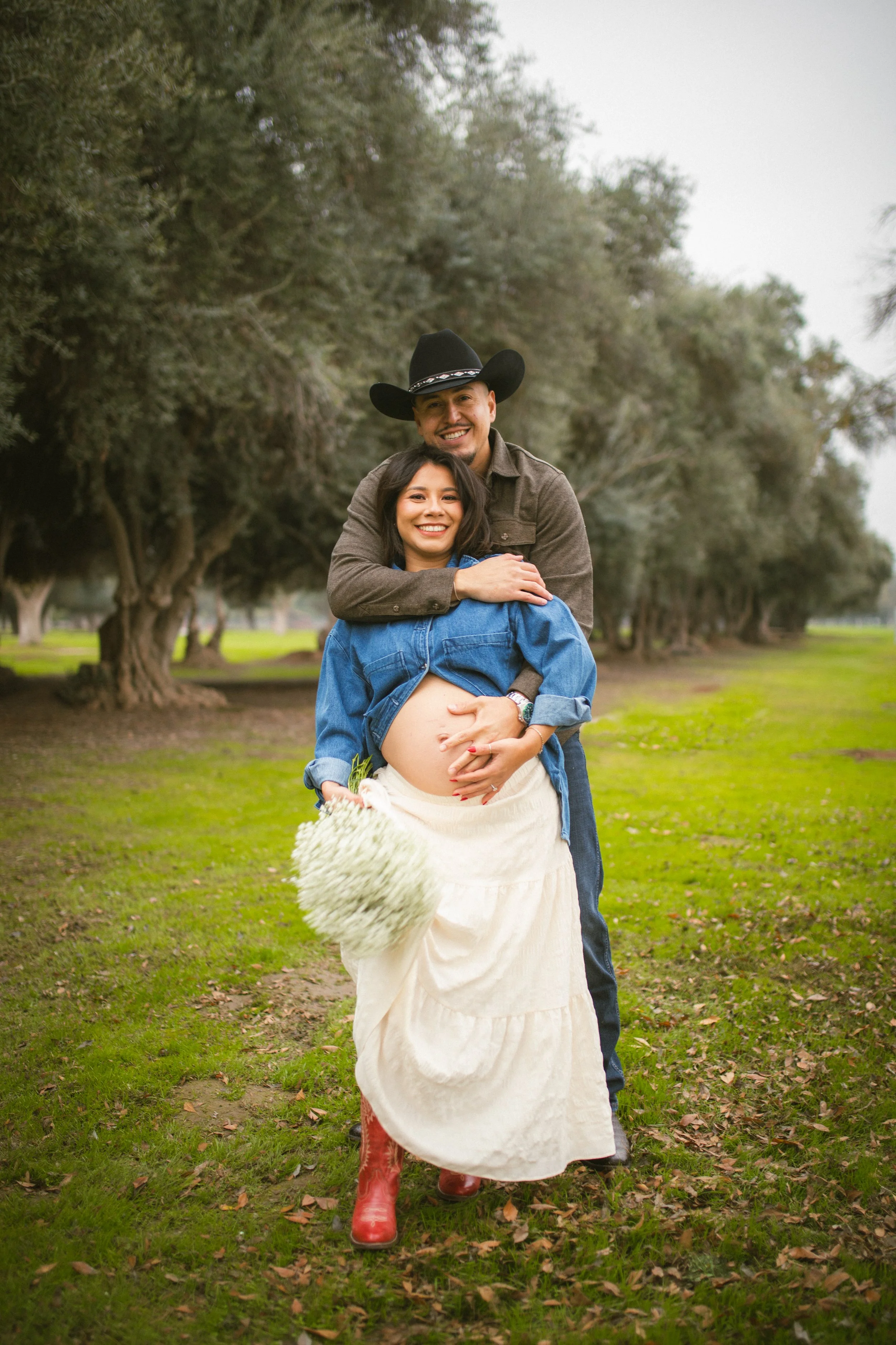 A smiling man wearing a cowboy hat and a woman with long dark hair, wearing a denim jacket and a white skirt, standing outside in a park with green grass and large trees. The woman is pregnant and holding a bouquet of white flowers, while the man is 